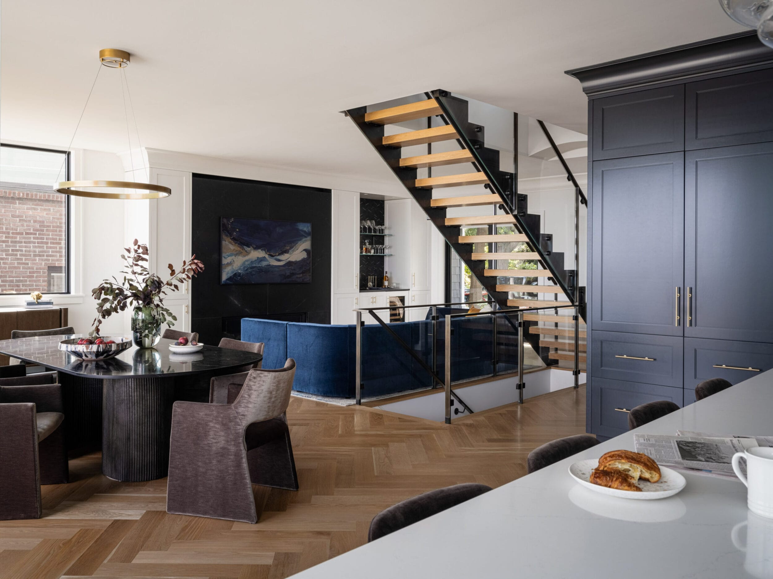 Modern open-concept living space with a dining area, stairway, and lounge seating. A croissant on a plate is on the kitchen counter.