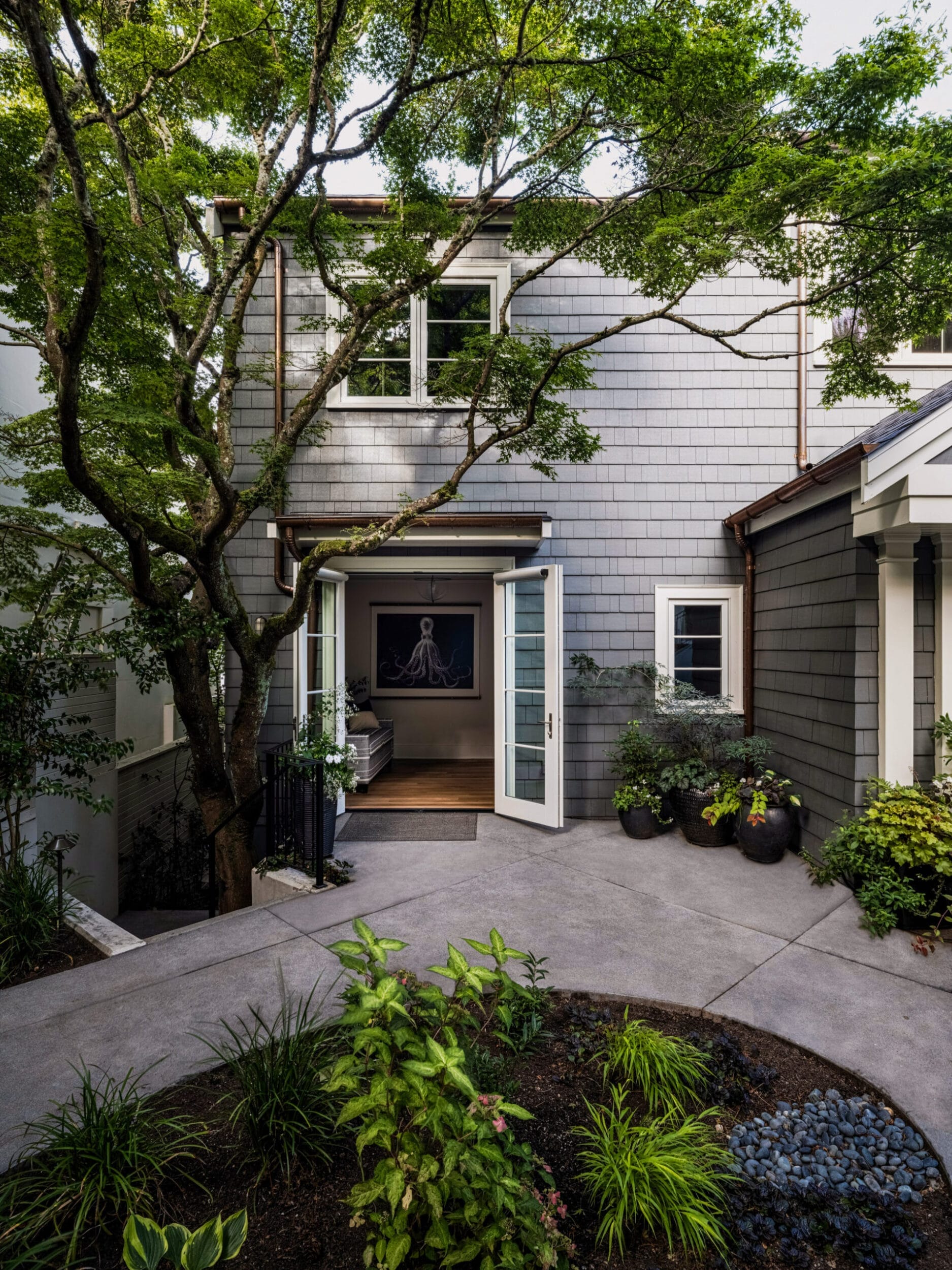 A modern house with gray siding, open French doors, and lush greenery in the foreground. A small tree is near the entrance, and a visible interior with artwork can be seen through the doors.