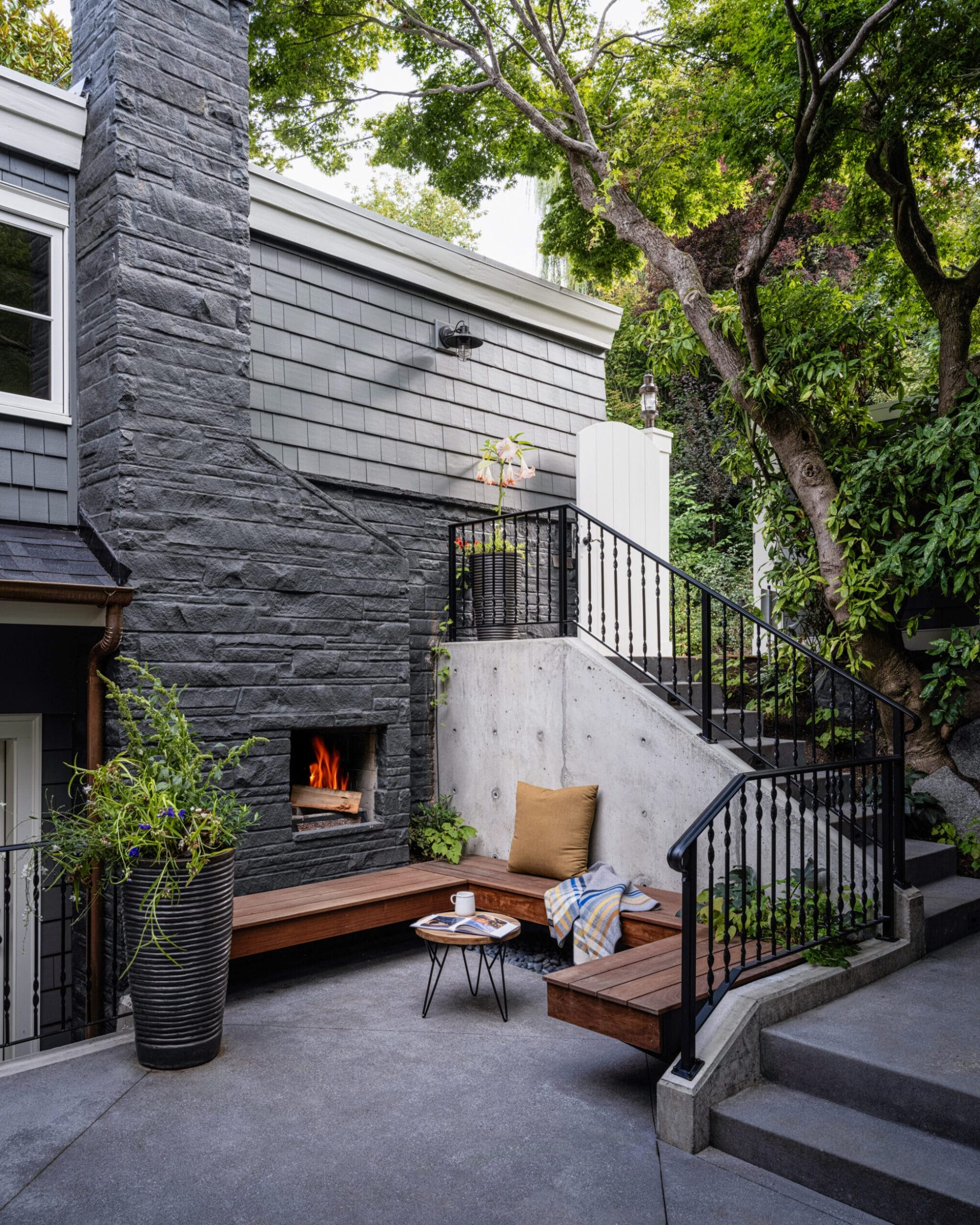 Outdoor patio with a stone fireplace, wooden seating, and potted plants. A cozy area with a small table and a blanket. Stairs lead up alongside a tree.