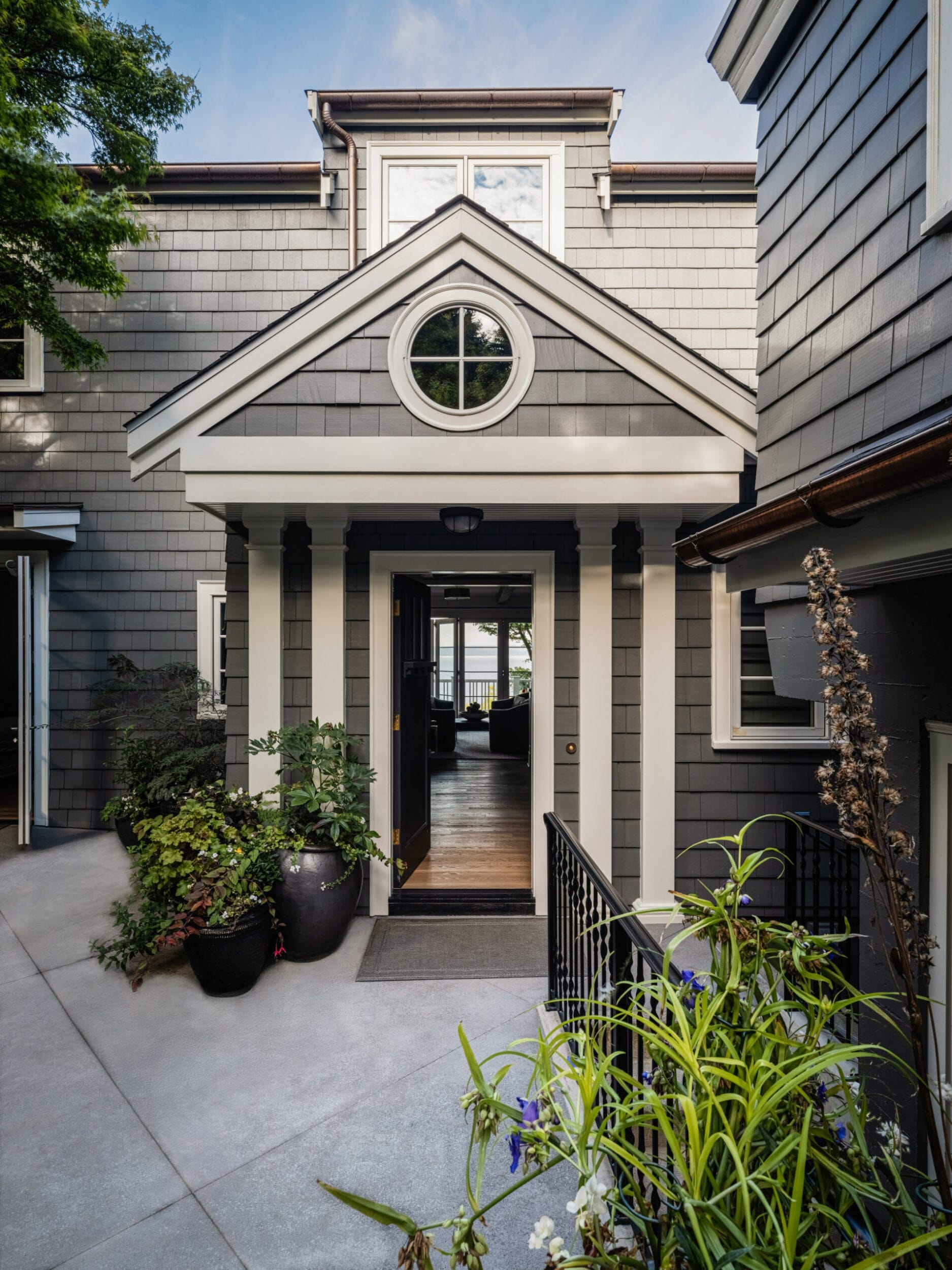 Entrance of a two-story gray house with white trim, featuring a gabled roof, circular window, and open door leading inside. Surrounding are plants and a concrete pathway.