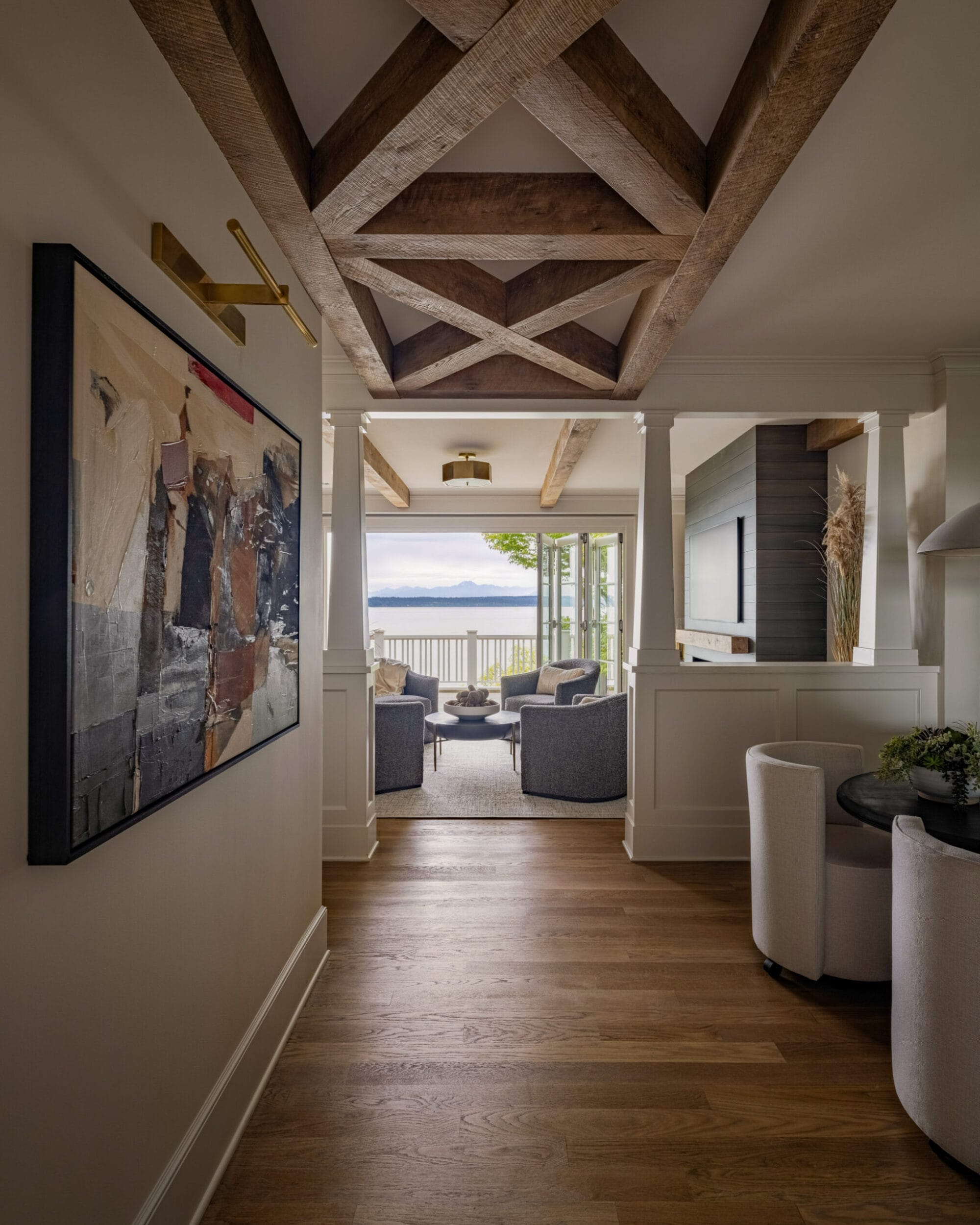 Hallway with wooden beams, a large abstract painting, and a view leading to a seating area with chairs and a table. Beyond, a balcony overlooks a scenic water view.