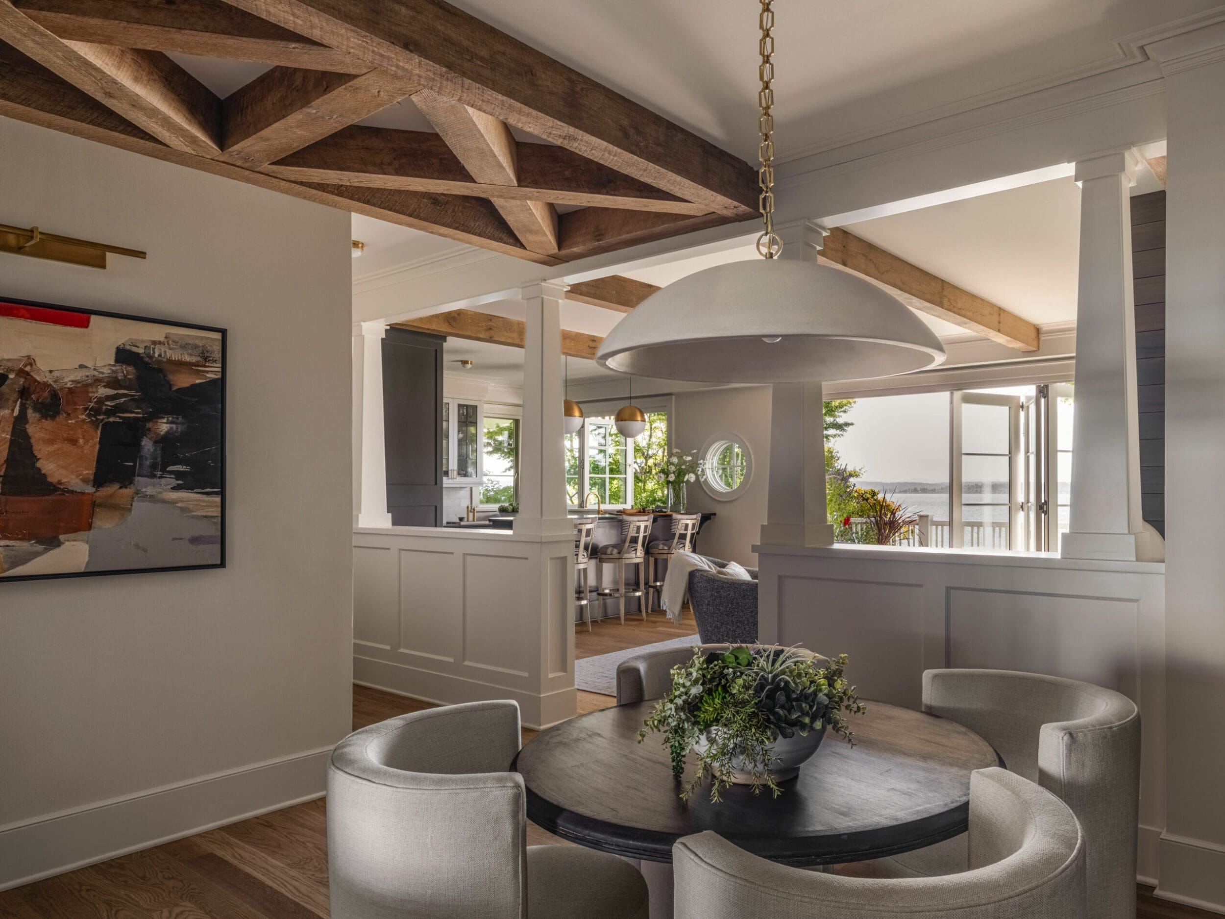 A dining area with a round table and four chairs. The room features exposed wooden beams, modern art on the wall, and a view of the outdoors through large windows.