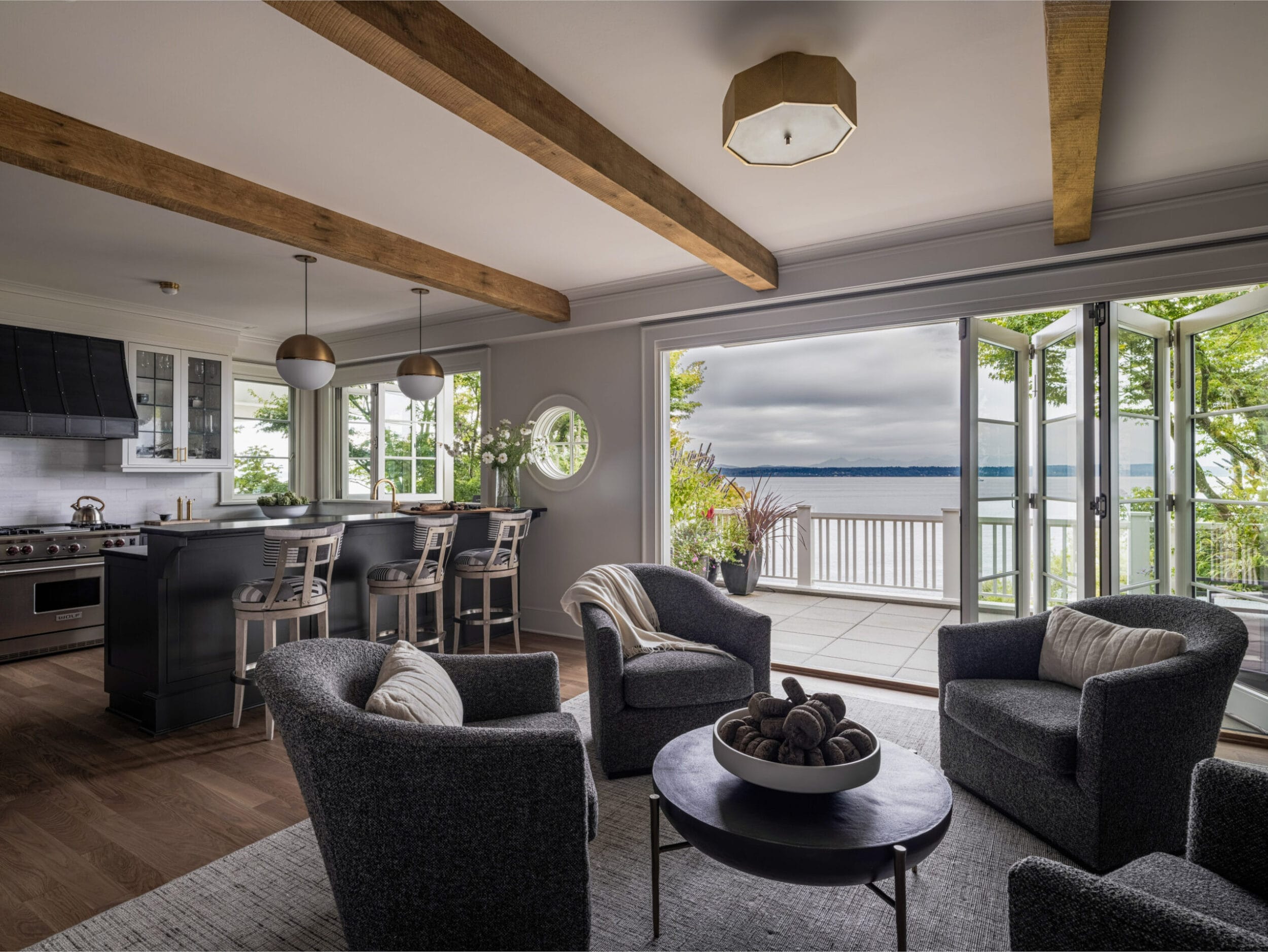 Modern open-plan living room and kitchen with gray furniture, wooden beams, and large windows opening to a balcony with a lake view.