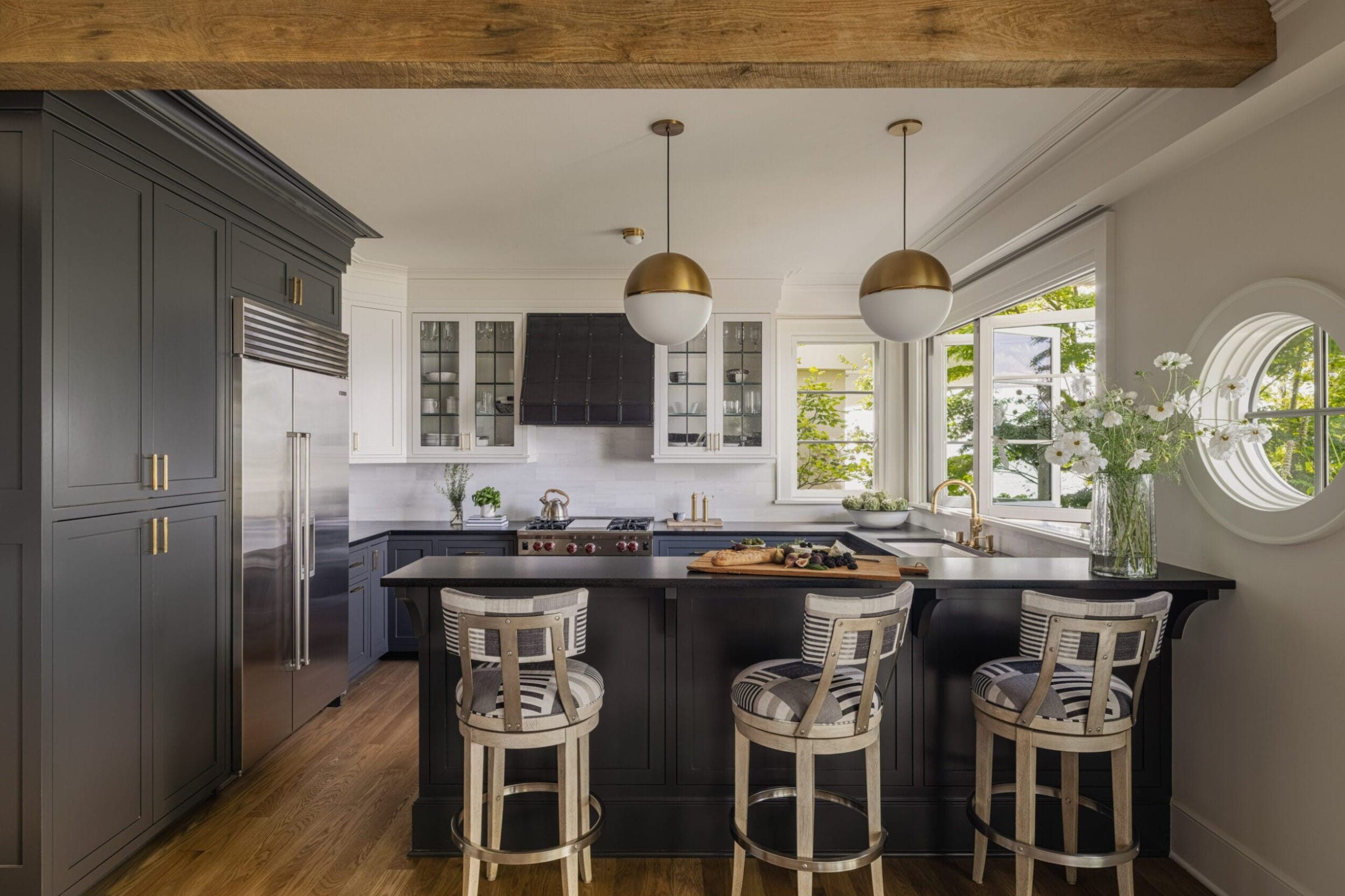 Modern kitchen with dark cabinetry, island with bar stools, pendant lights, and stainless steel appliances. Large window and decorative beams enhance the space.