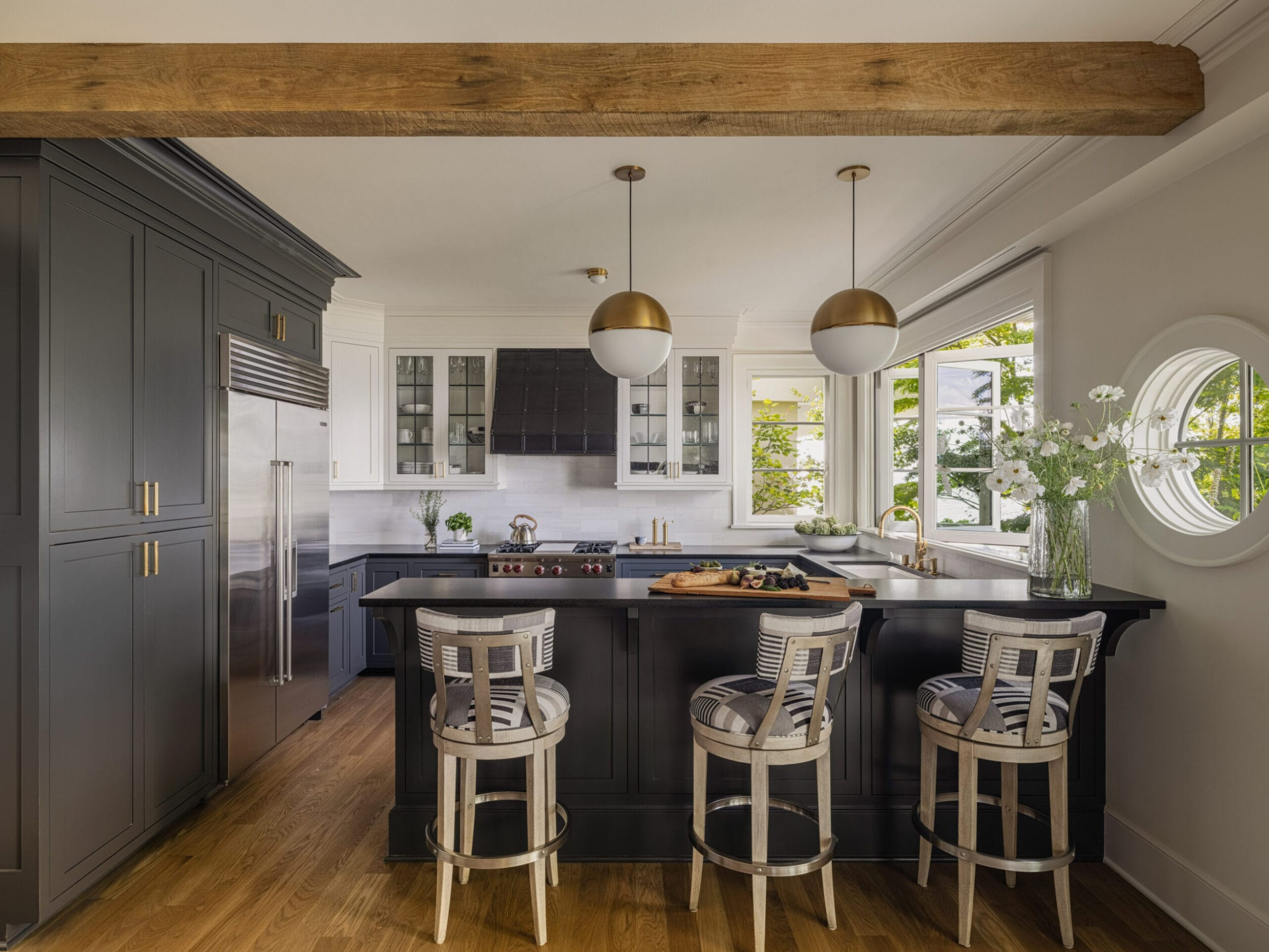 Modern kitchen with dark cabinetry, island with bar stools, pendant lights, and stainless steel appliances. Large window and decorative beams enhance the space.