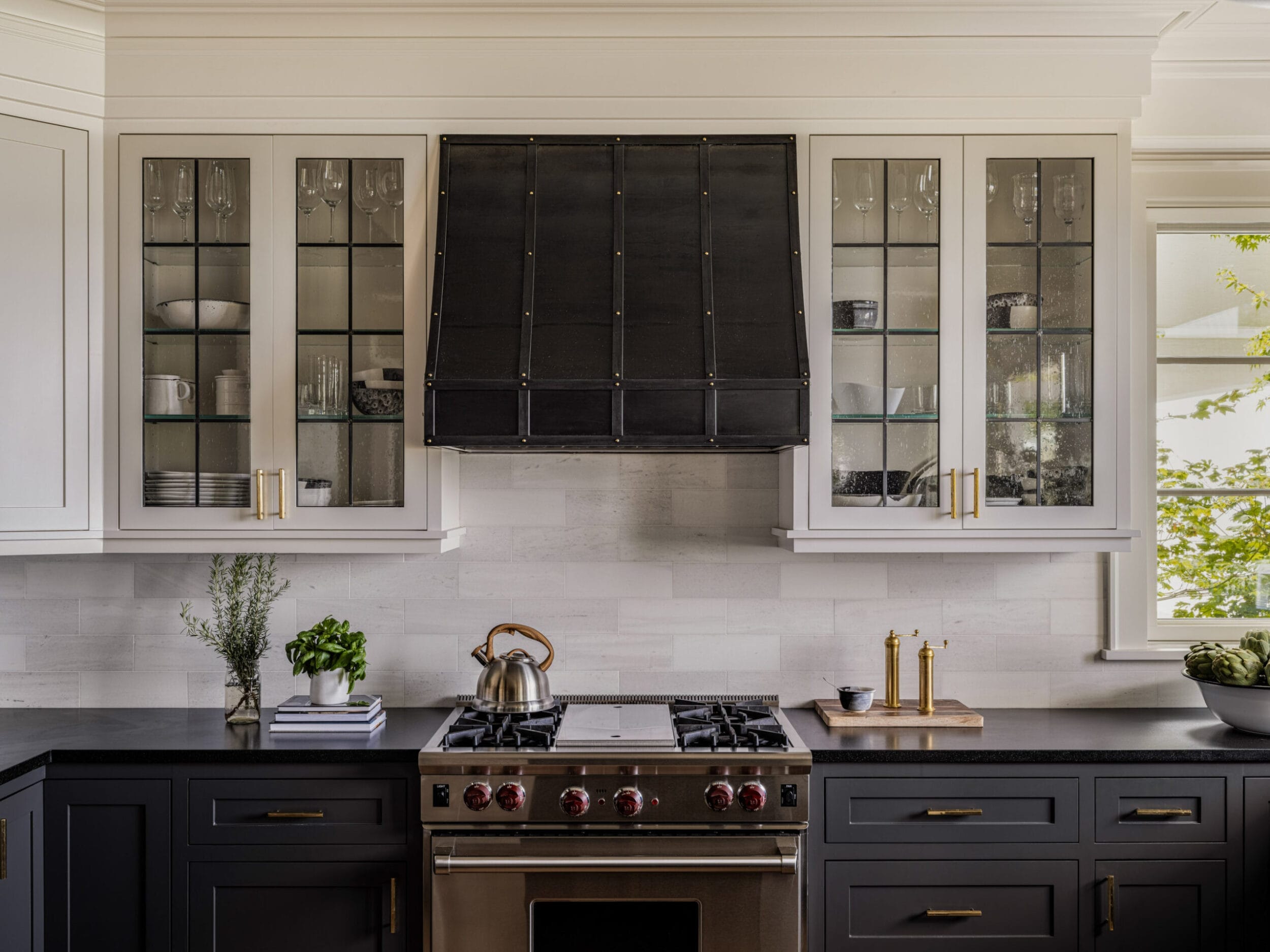 Modern kitchen with a black stove and hood, white cabinets with glass doors, black countertop, and various kitchen items like a kettle, plants, and a bowl of apples.