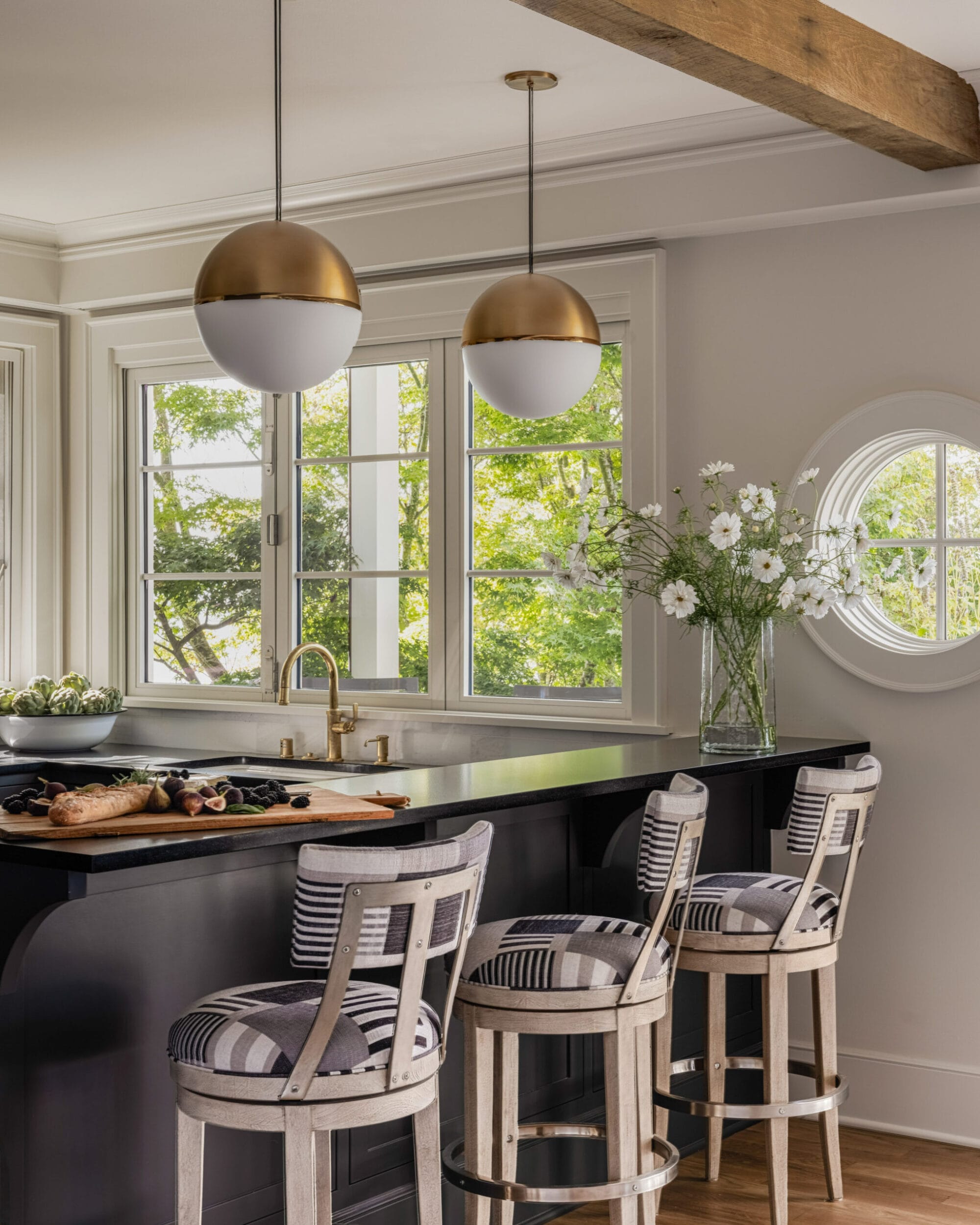 A modern kitchen with a black island, striped stools, and brass pendant lights. Large windows and a vase with white flowers add brightness to the space.