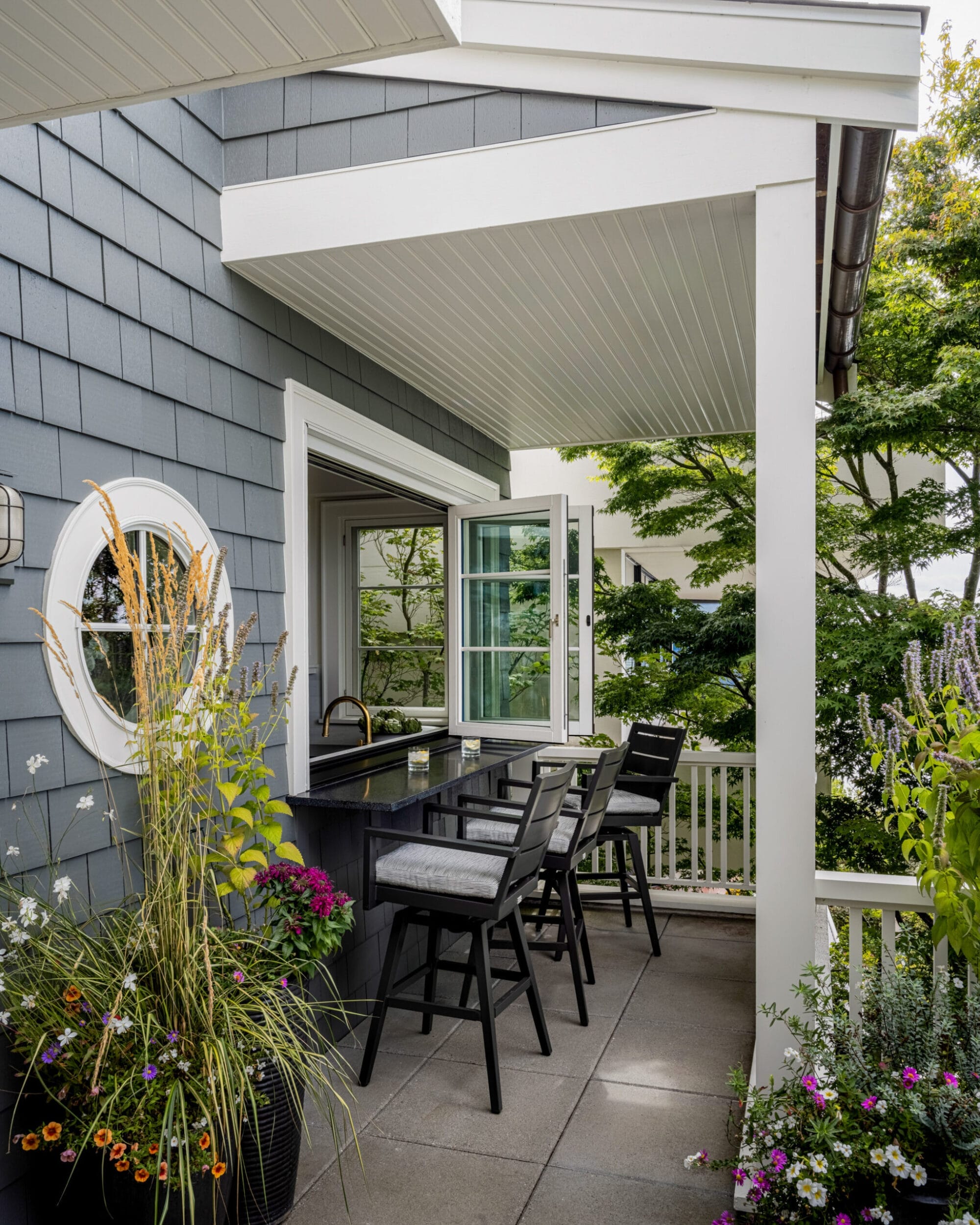 A small outdoor patio with a bar-height counter and chairs, surrounded by potted plants and flowers, adjacent to a house with blue-gray siding and an open window.