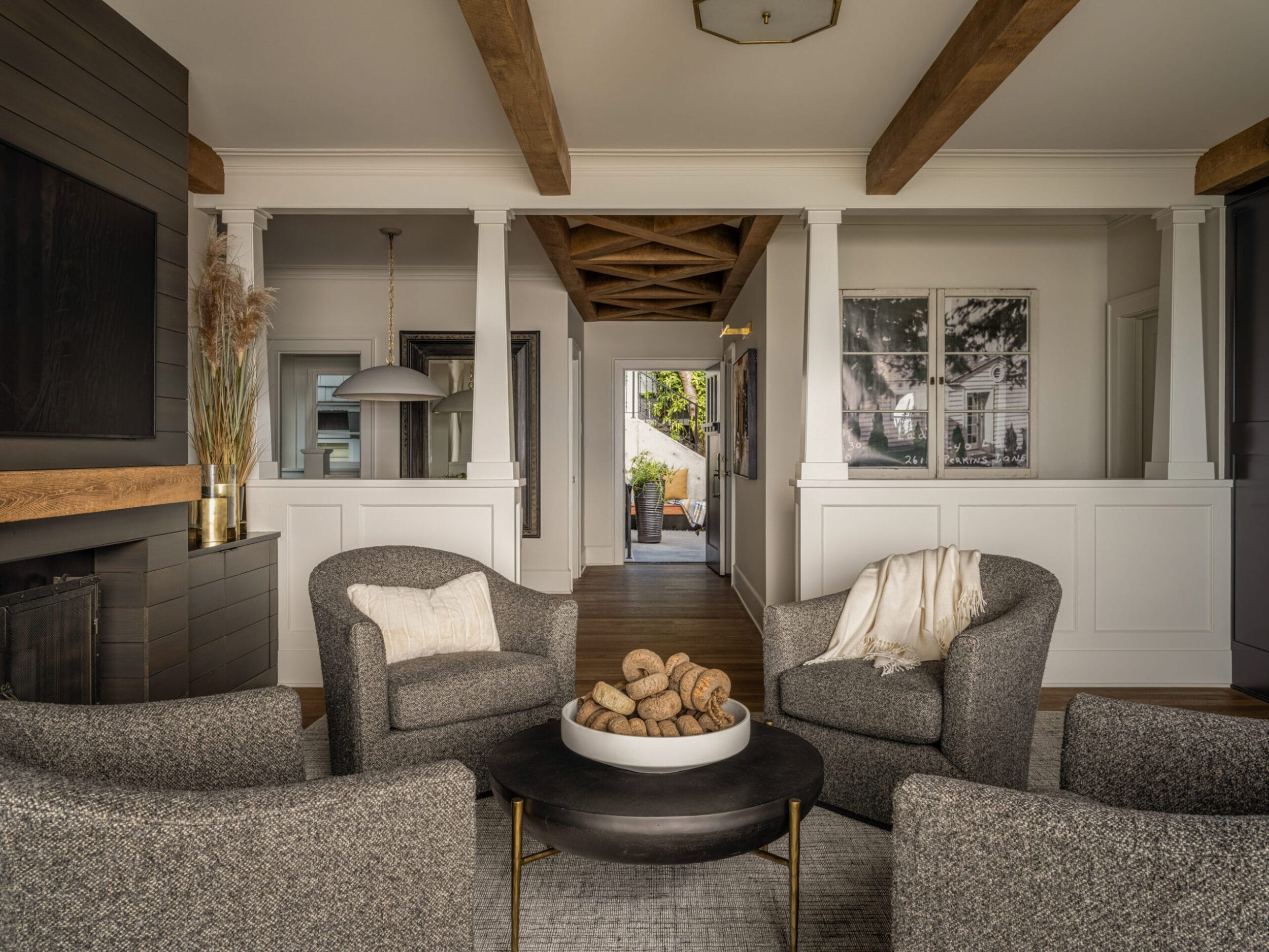 A cozy living room with gray armchairs, a round coffee table with decorative items, a fireplace, and a view of a hallway leading outside. The ceiling features exposed wooden beams.