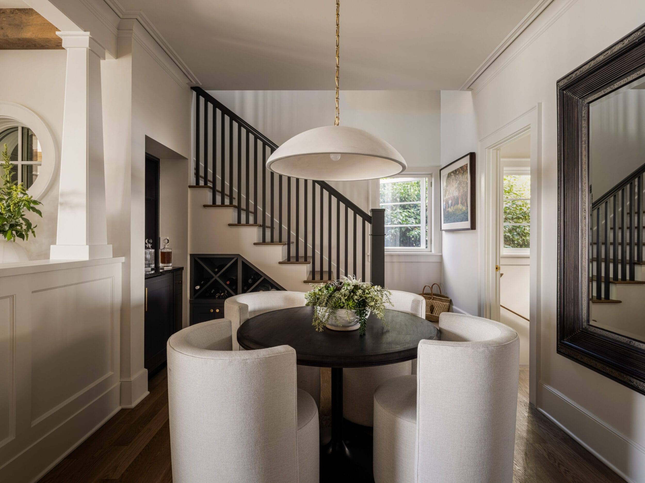 A dining area with a round table and four chairs. A large overhead light hangs above. A staircase and a mirror adorn the room. A window provides natural light.