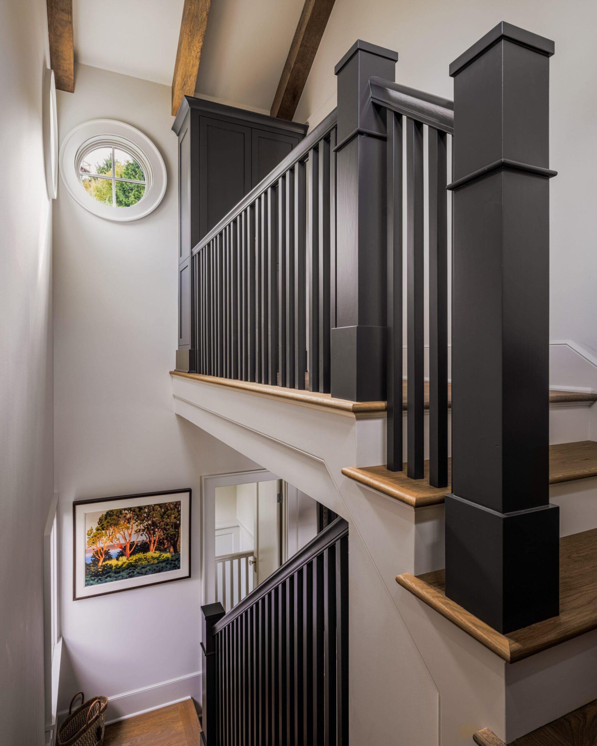 A staircase with black railings, wooden steps, and a round window at the top. A framed picture hangs on the wall.