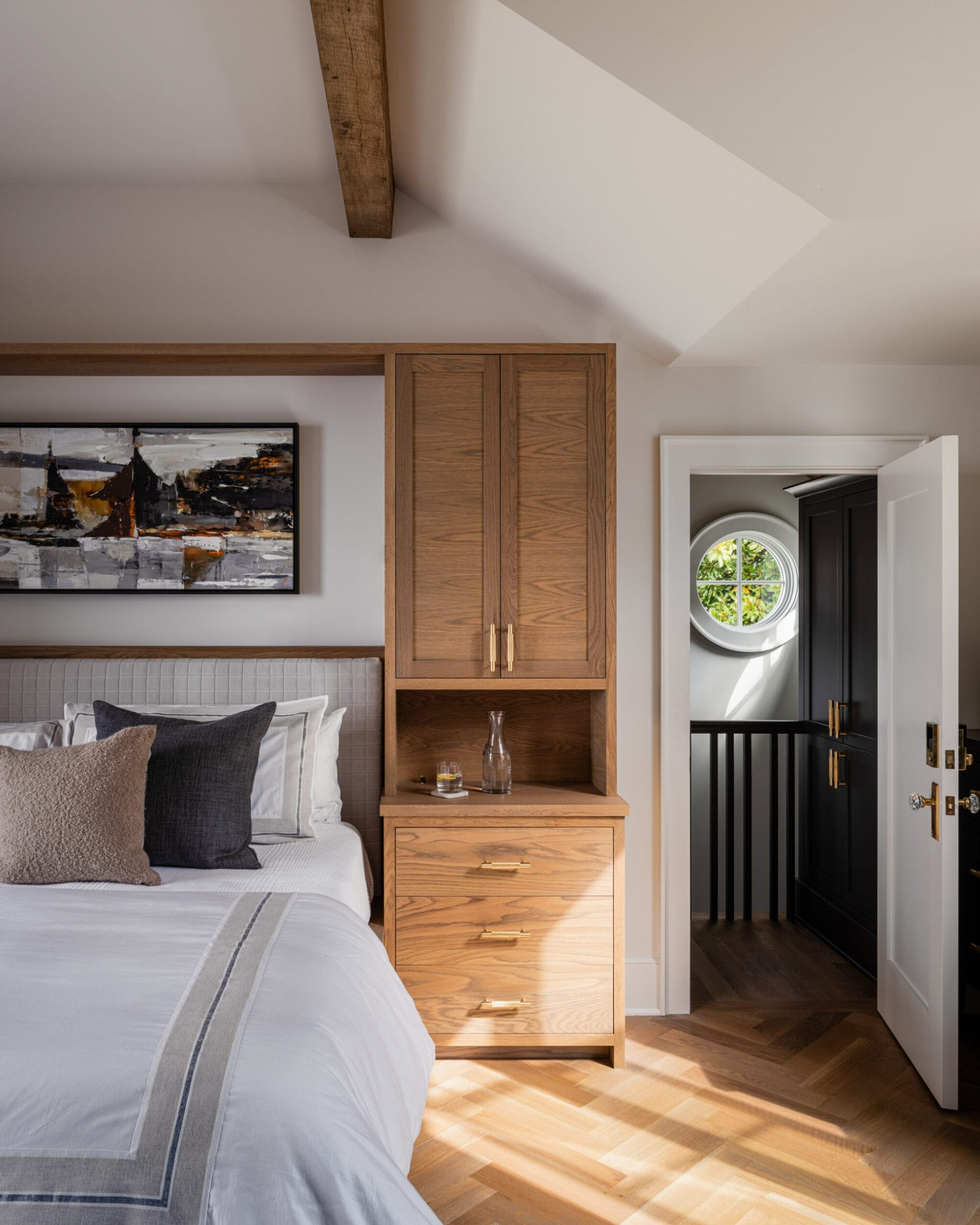 Modern bedroom with a bed, wooden nightstand, and abstract wall art. A door on the right opens to a hallway with a round window. Exposed beam ceiling adds a rustic touch.