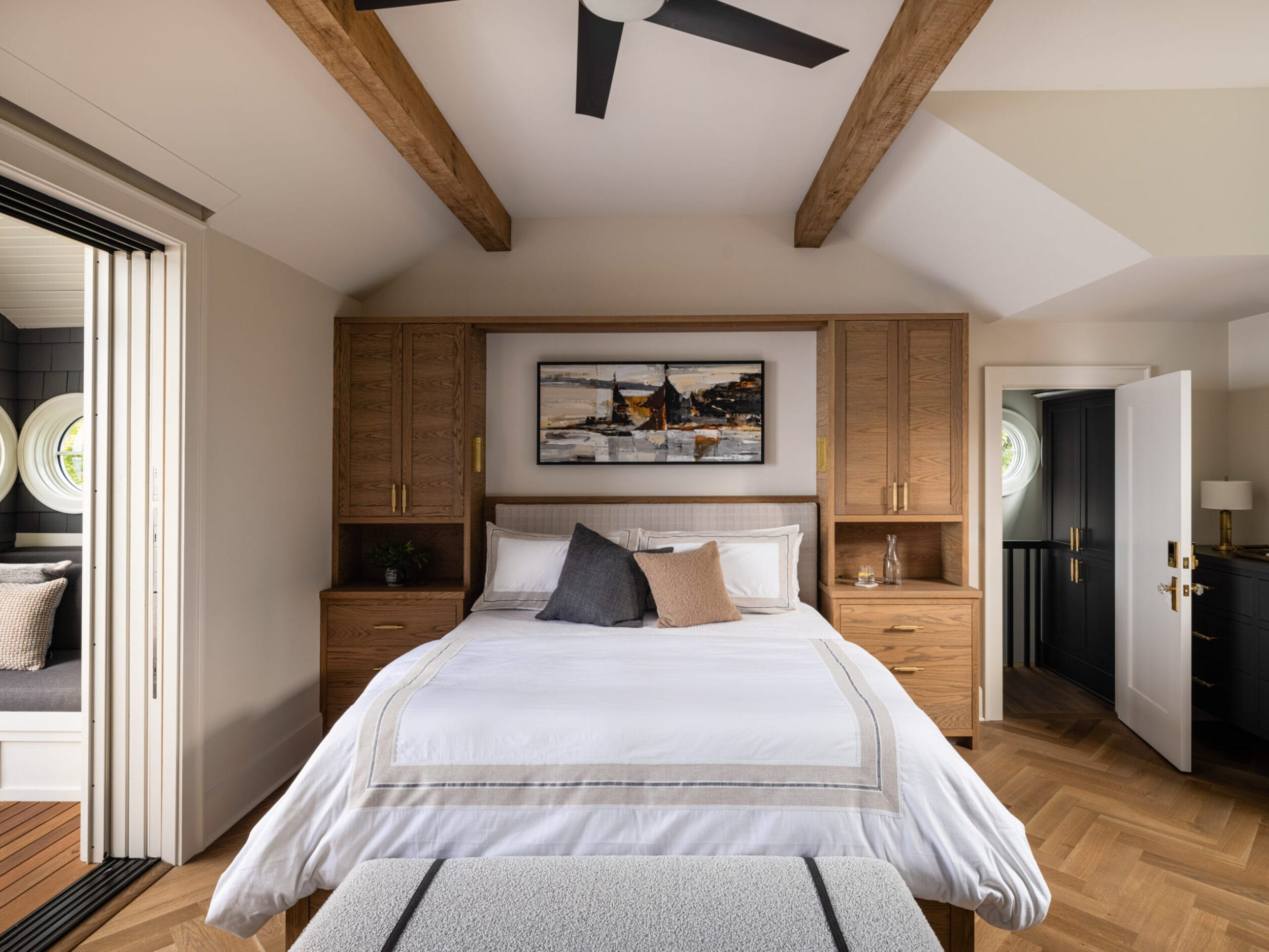 Bedroom with a large bed, wooden headboard, and ceiling beams. Artwork hangs above the bed. A sliding door opens to an outdoor space on the left. A doorway leads out on the right.