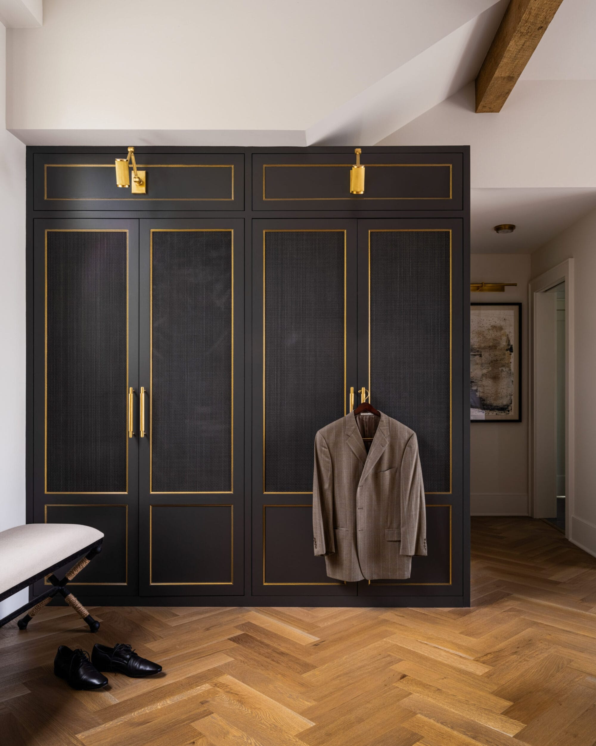 A gray blazer hangs on black wardrobe doors with gold accents in a modern room featuring herringbone wood flooring, a bench, and a pair of dress shoes.