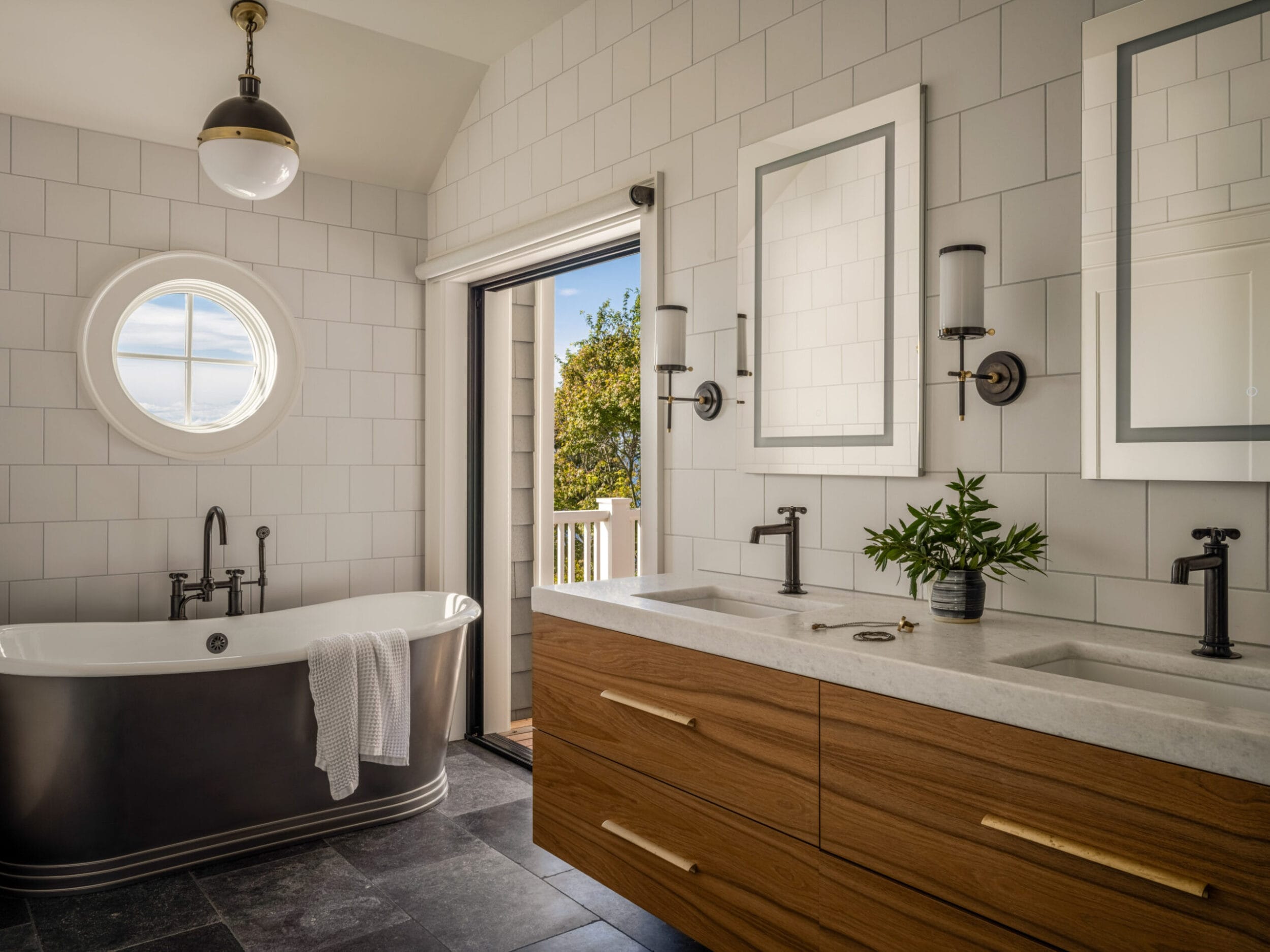 Modern bathroom with freestanding tub, double vanity, wall-mounted mirrors, circular window, and open door leading to a balcony with trees visible outside.