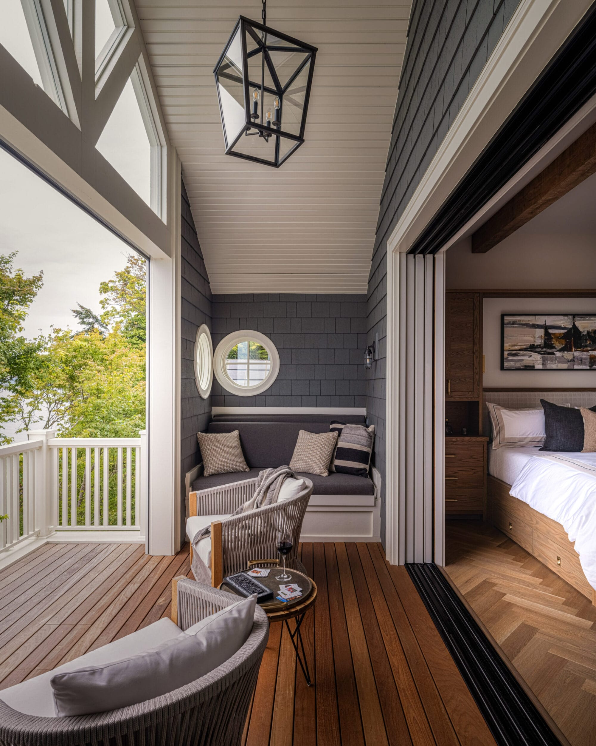 A cozy, modern balcony with wooden flooring, two chairs, a bench, and a hanging light fixture, connected to a bedroom. Lush greenery is visible outside.