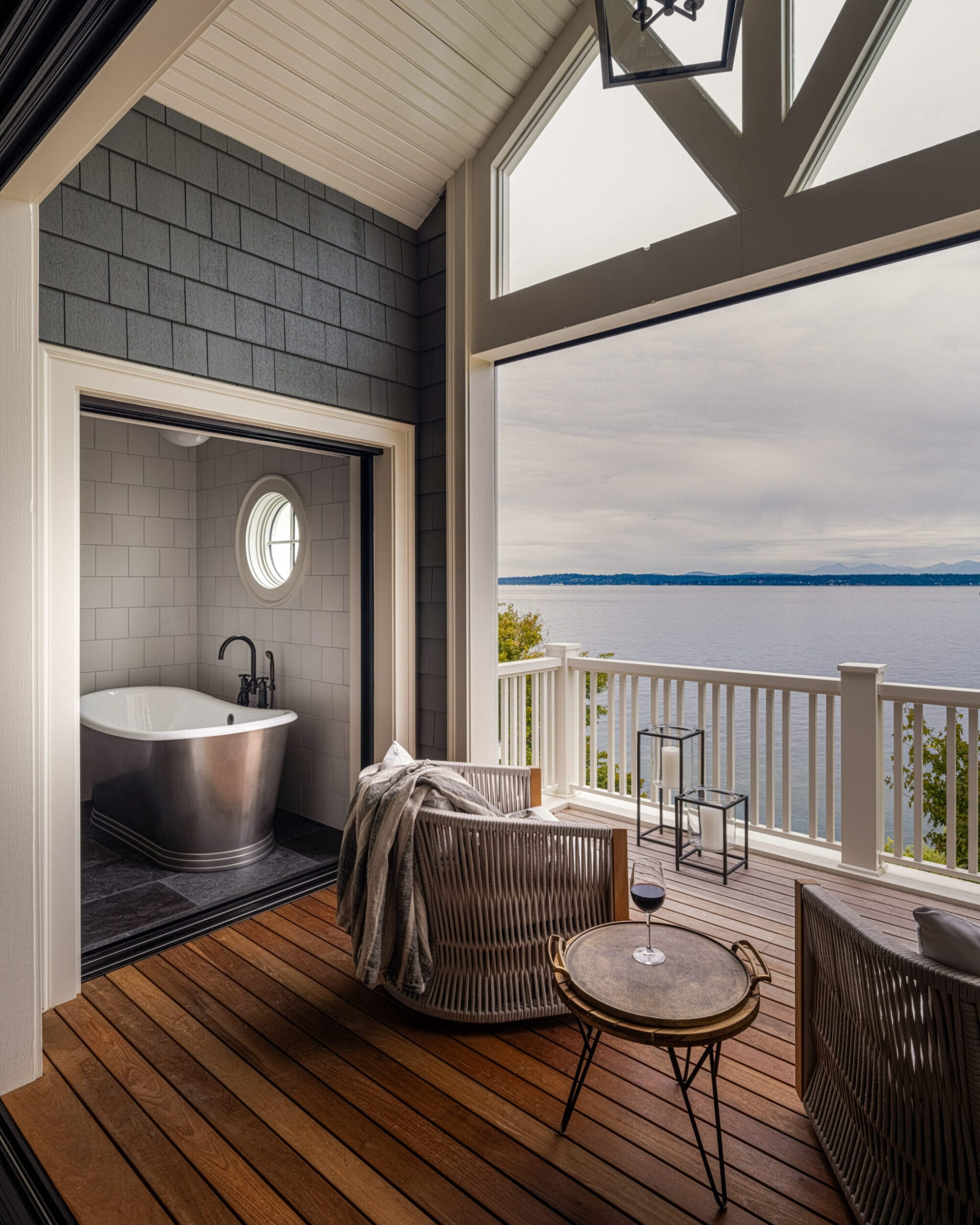 A cozy deck with wicker seating overlooks a lake. The interior features a bathtub with a circular window. A small table holds a wine glass, and a light fixture hangs from the ceiling.