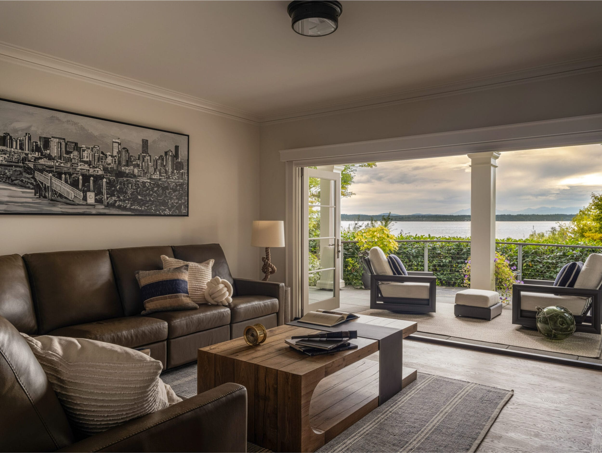 A living room with a brown sofa, a wooden coffee table, and a cityscape painting on the wall. Large glass doors open to a patio with a stunning view of the water and sky.