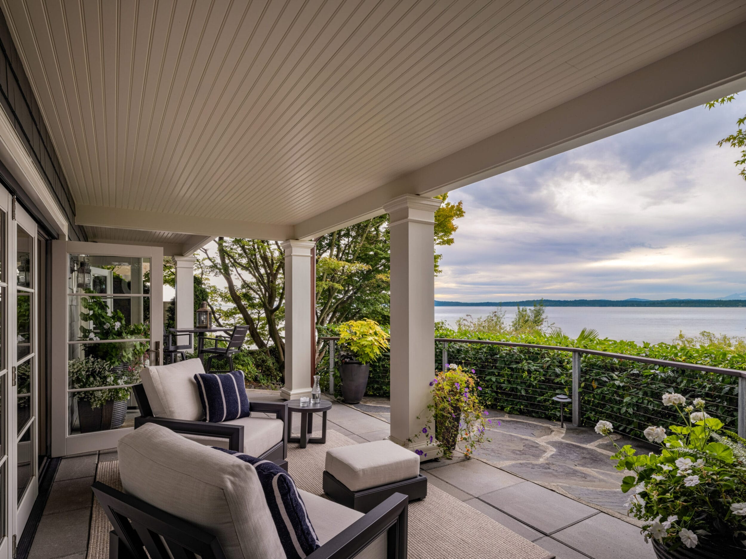 Covered patio with white cushioned chairs overlooking a scenic body of water. Surrounded by leafy plants and greenery, with overcast sky in the background.