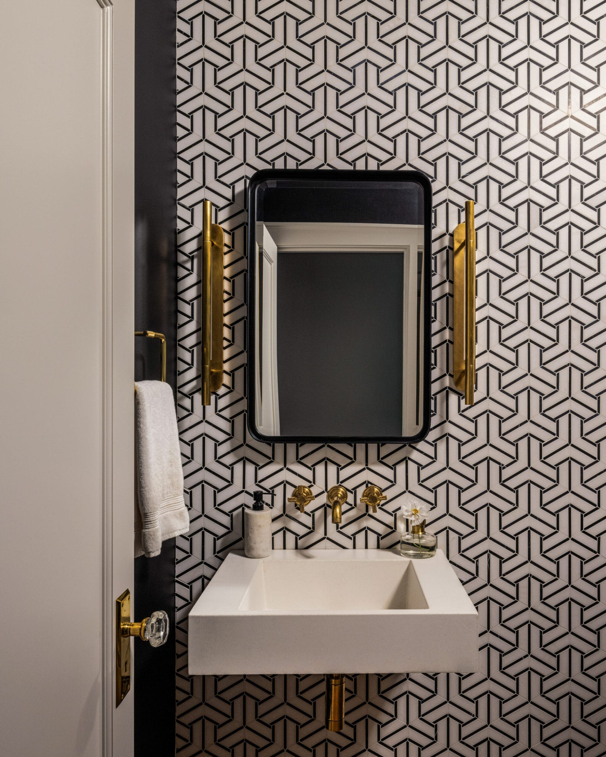 Bathroom with geometric patterned wallpaper, a rectangular mirror, gold faucets, wall sconces, and a white rectangular sink.