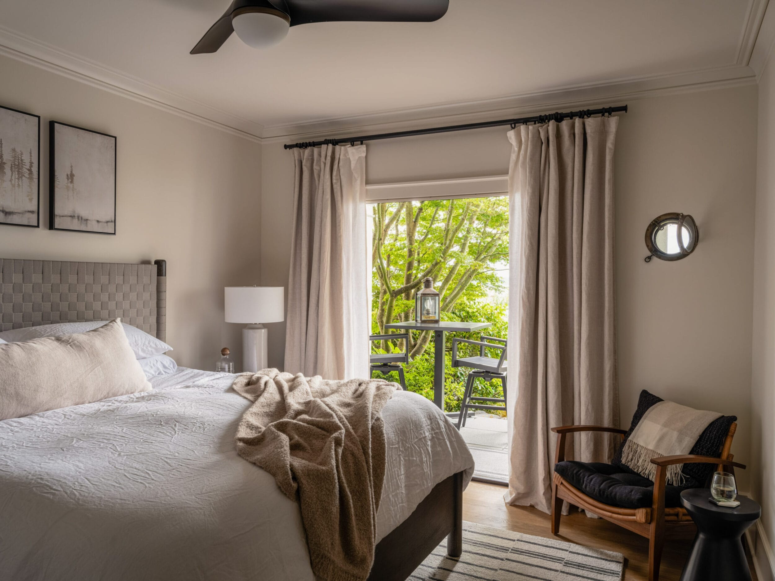 Bedroom with a neatly made bed, a chair, and framed art on the wall. A sliding glass door leads to a patio with green foliage visible outside.