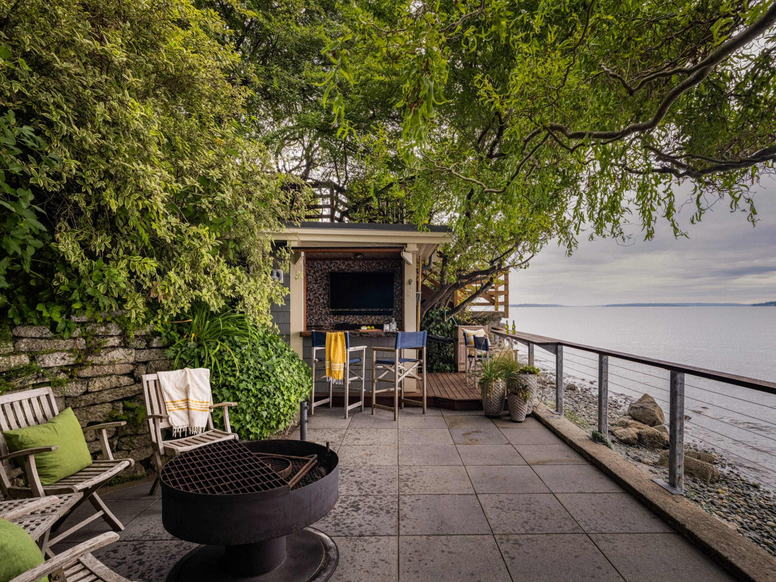 Outdoor seating area by the water with chairs, a fire pit, and a wall-mounted television under lush trees.