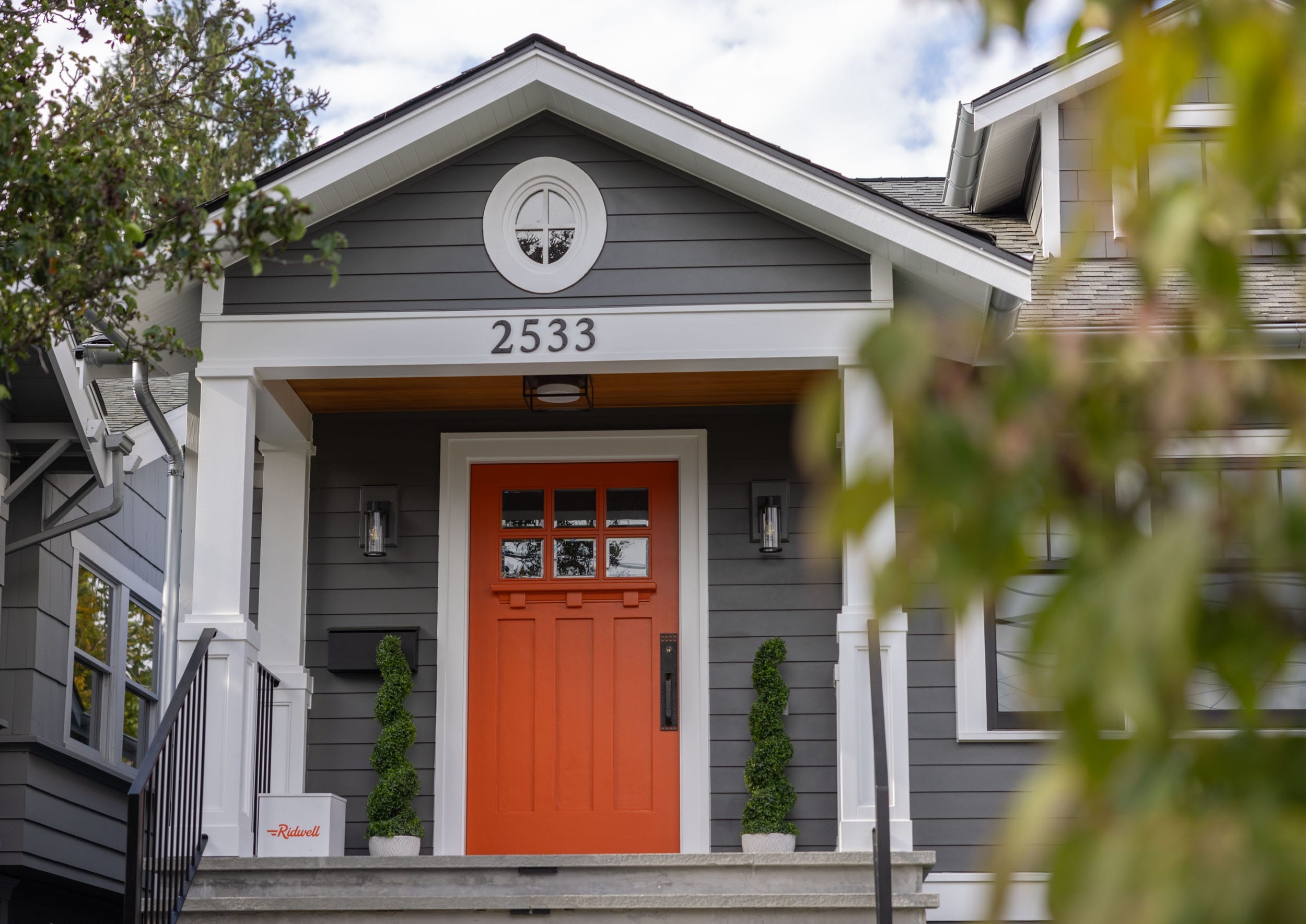 A gray house with a bright orange front door and the number 2533 above it. Two potted plants flank the entrance.