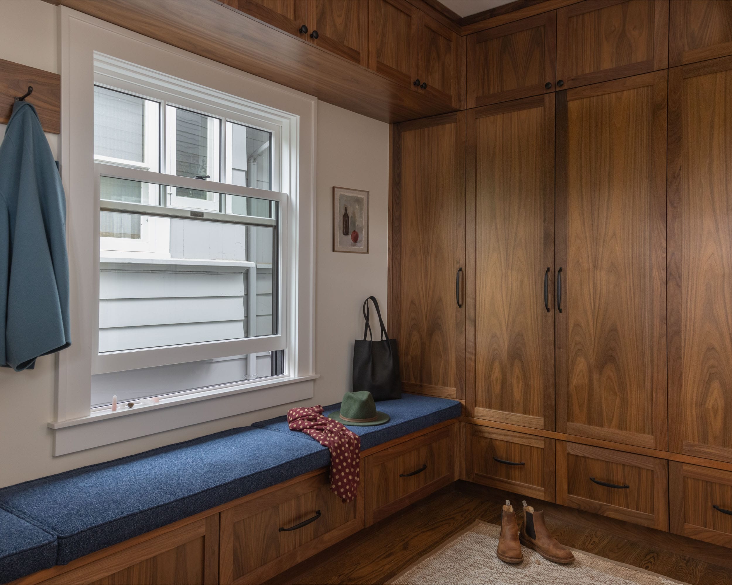 A cozy mudroom with built-in wooden cabinets and a blue cushioned bench under a window. A green hat, red scarf, black bag, and brown boots are placed on the bench and floor.