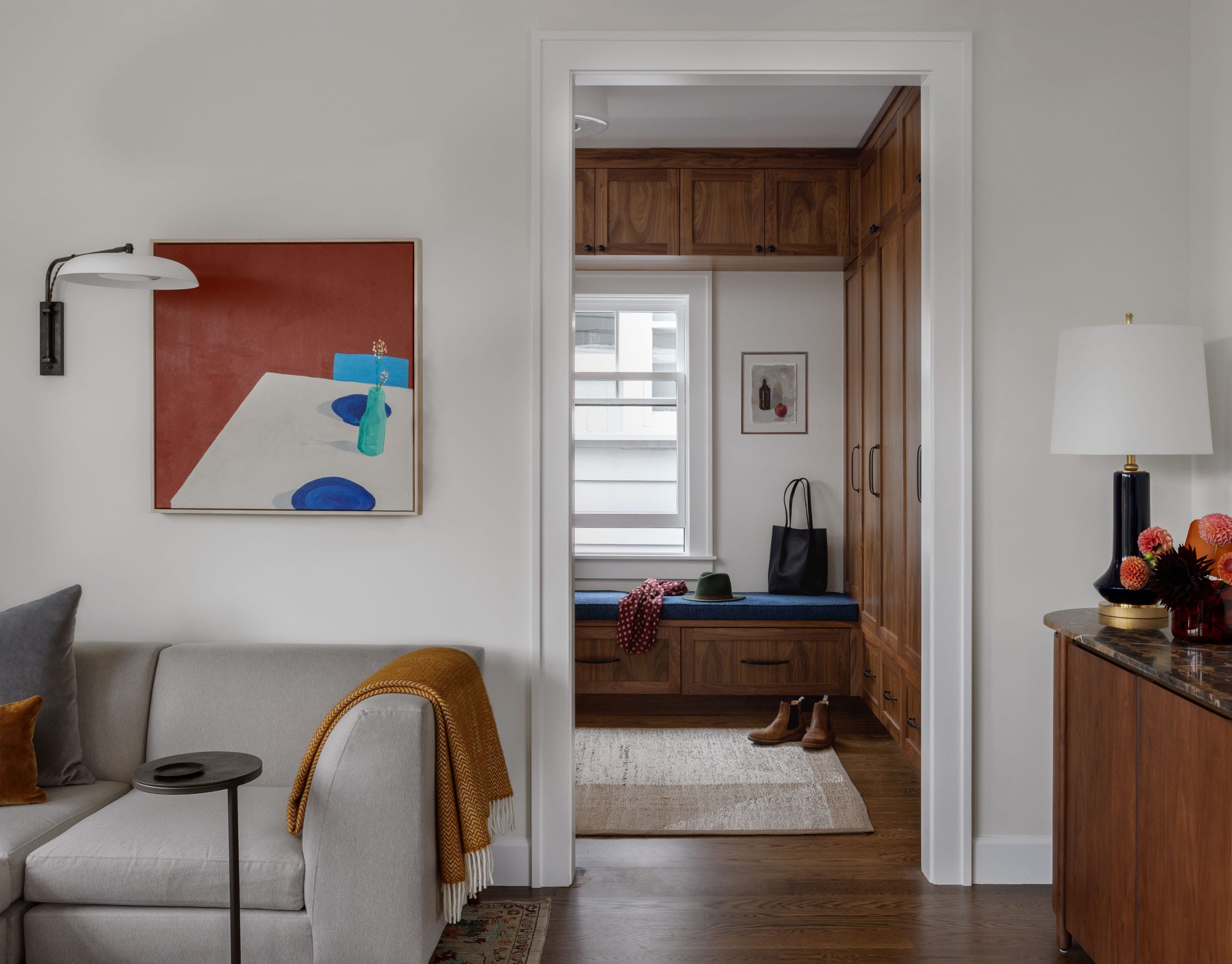 A cozy living room with a gray sofa, orange blanket, and modern artwork. A hallway leads to a window with a bench, wooden cabinets, shoes, and a bag. A sideboard with a lamp and flowers.