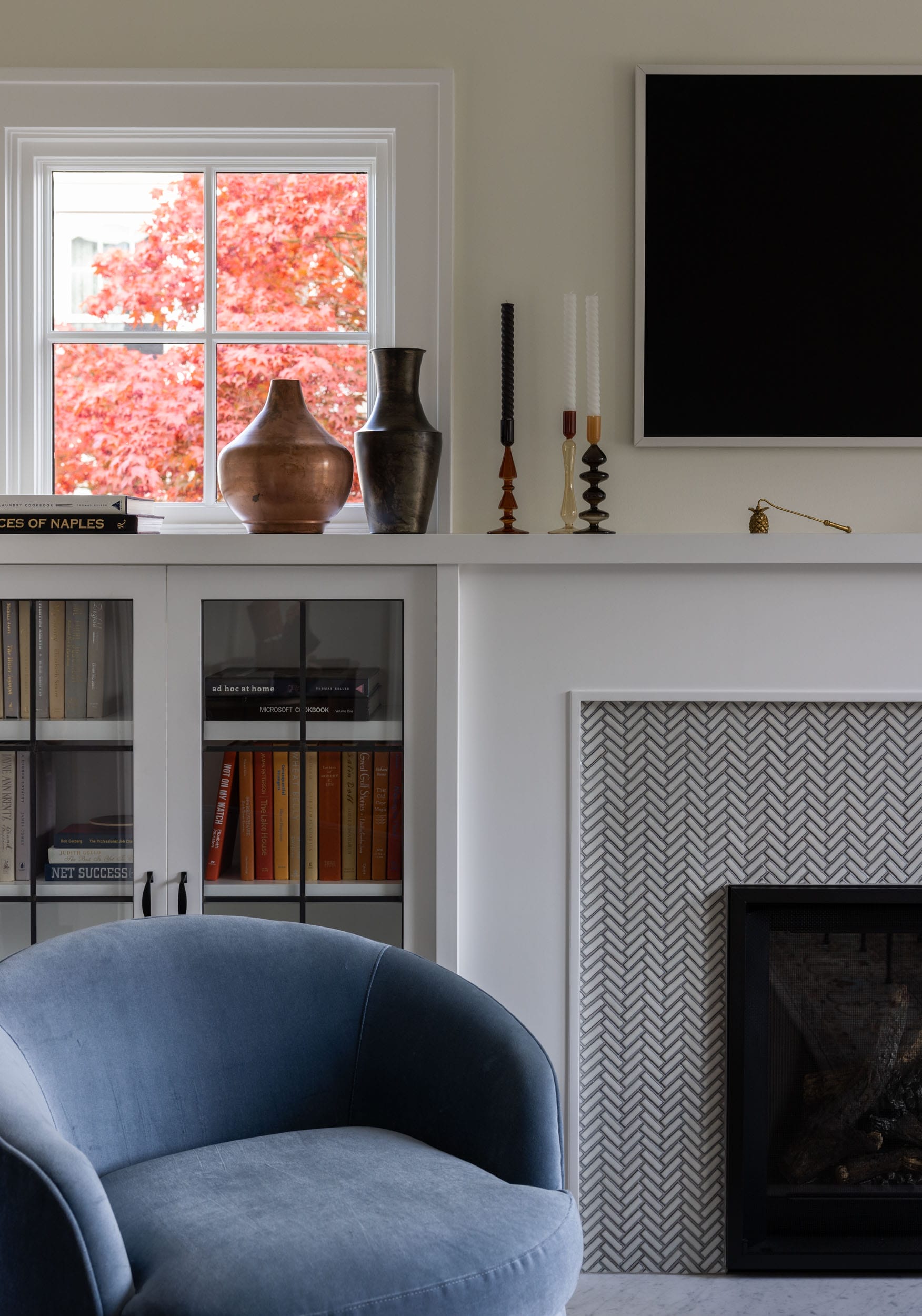 A modern living room features a blue chair, a white mantle with decorative vases and candles, a black framed TV, a built-in bookshelf, and a window with a view of red autumn leaves.