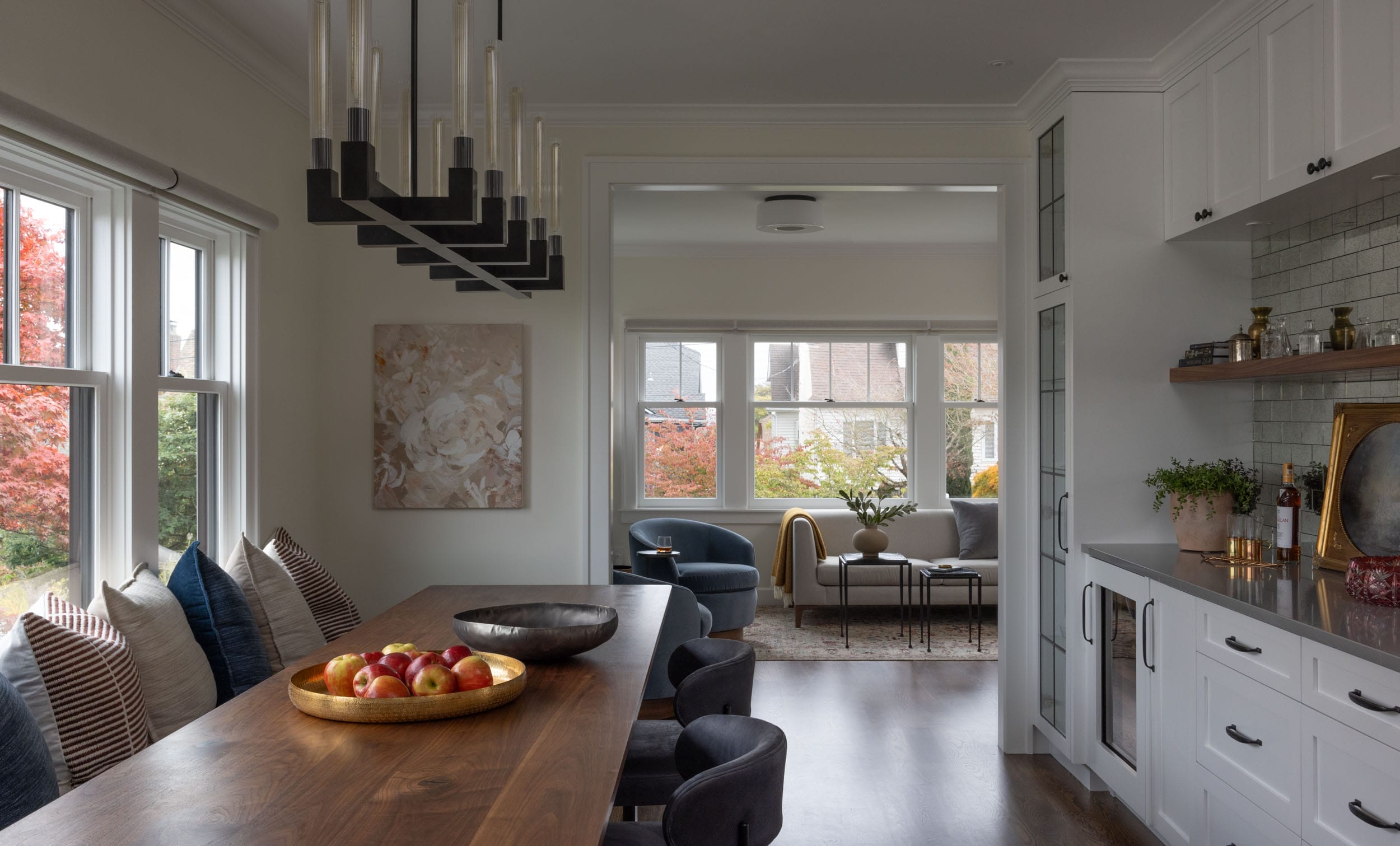 A dining area with a wooden table and a black chandelier. A bowl of apples is on the table with striped pillows on the chairs. In the background, a living room with large windows and armchairs.