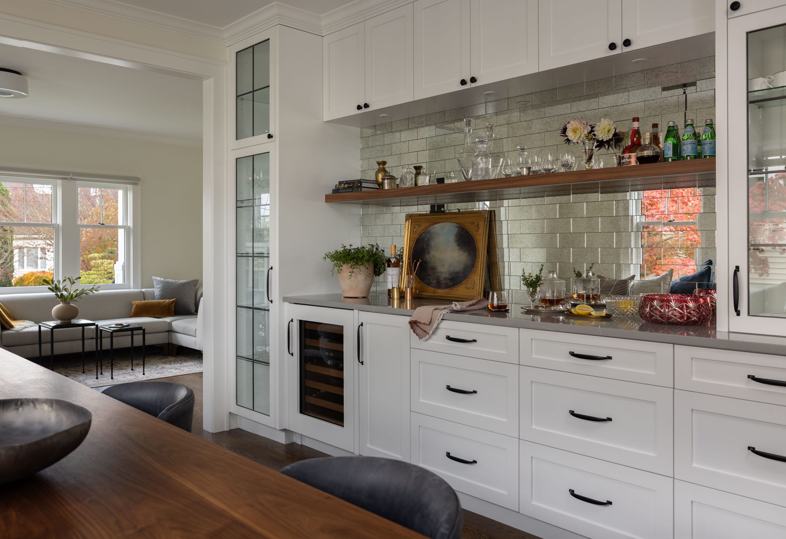 Modern kitchen with white cabinetry, mirrored backsplash, and a built-in wine cooler. A wooden countertop and decor items are displayed, with a glimpse of a cozy room and windows in the background.