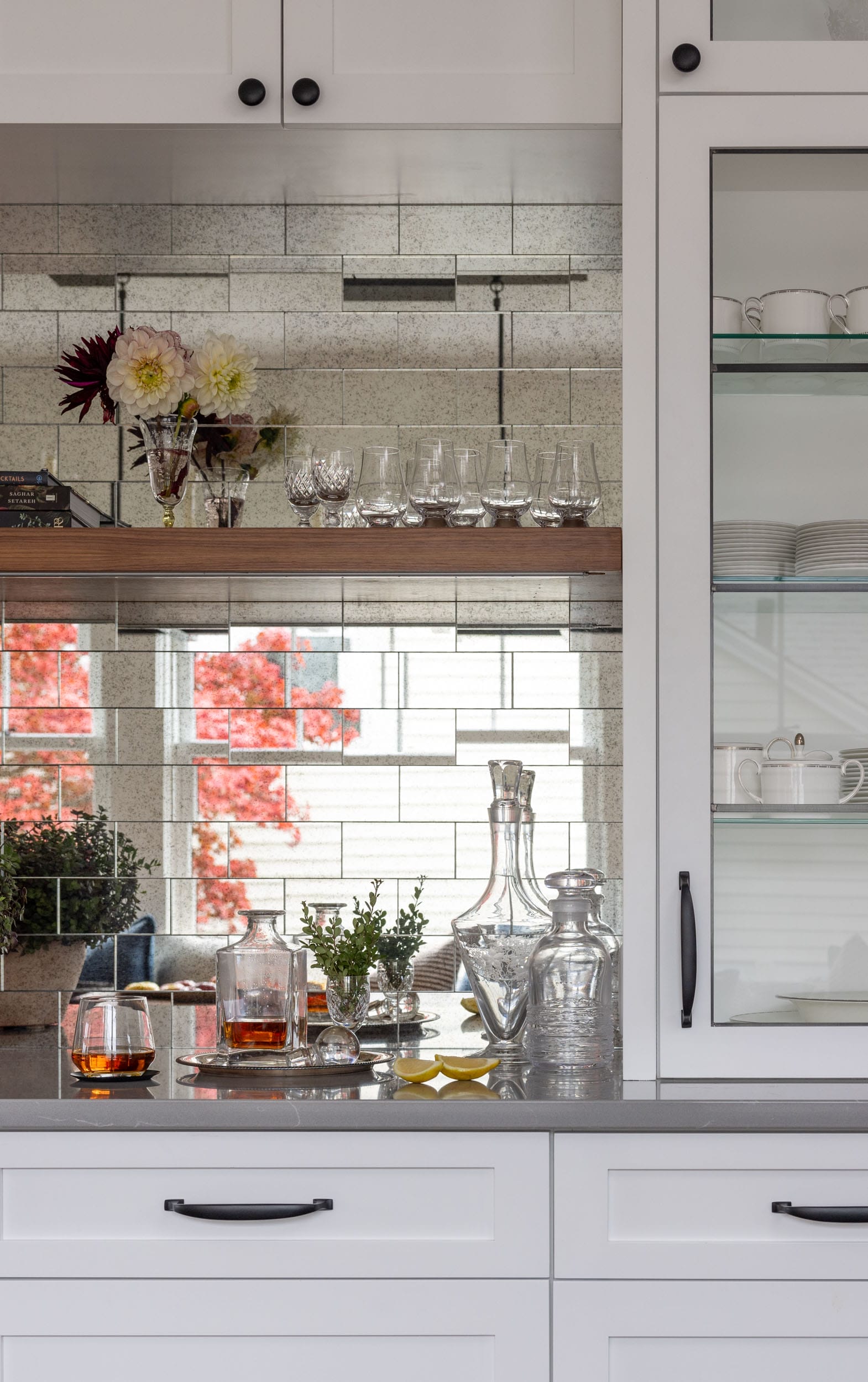 Modern kitchen bar area with mirrored backsplash, glassware on wooden shelf, and assorted glass decanters and tumblers on countertop. Red foliage visible in reflection.