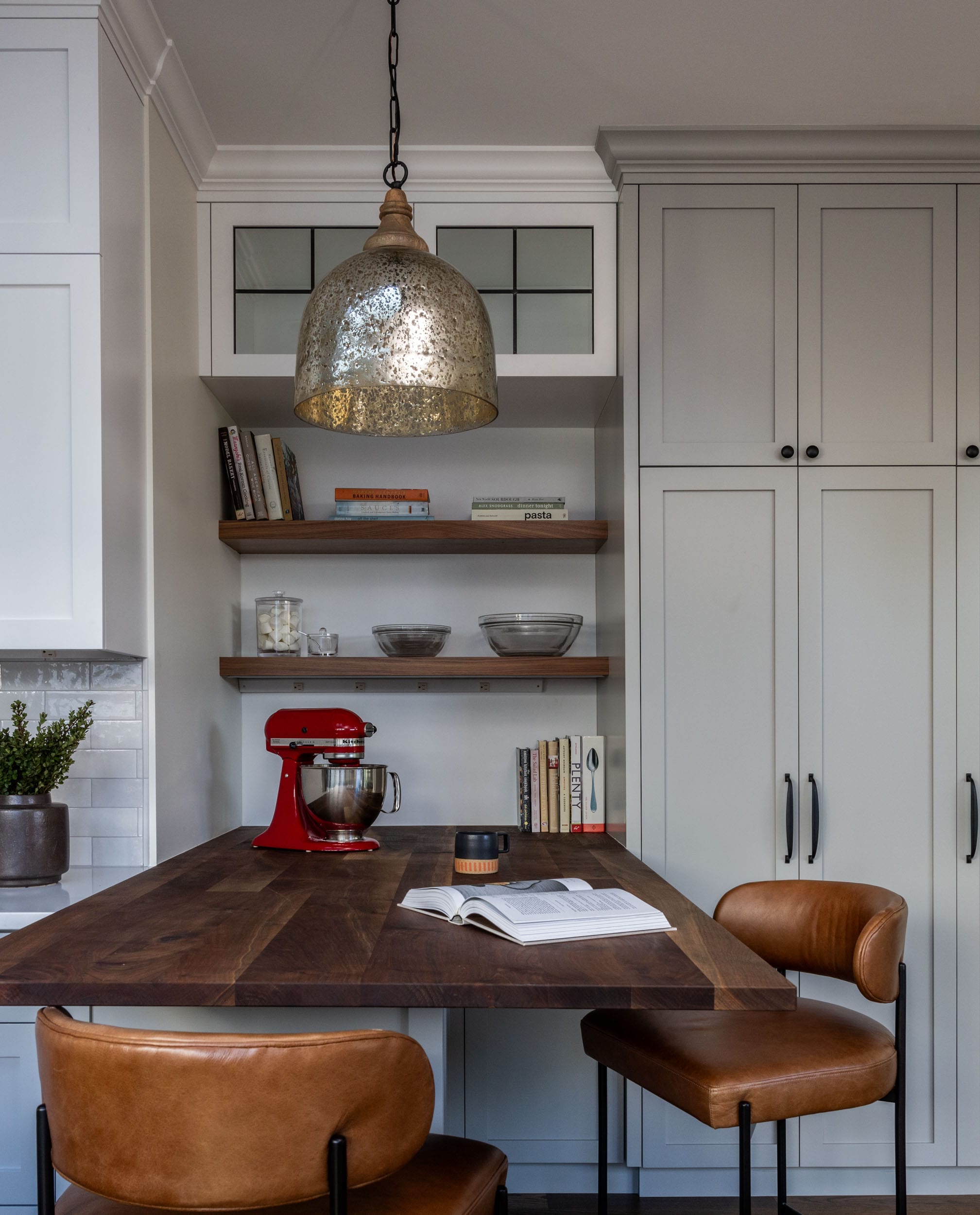 Modern kitchen with a wooden island, leather barstools, a hanging pendant light, and a red stand mixer. Shelves hold glass bowls and books. White cabinetry lines the wall.