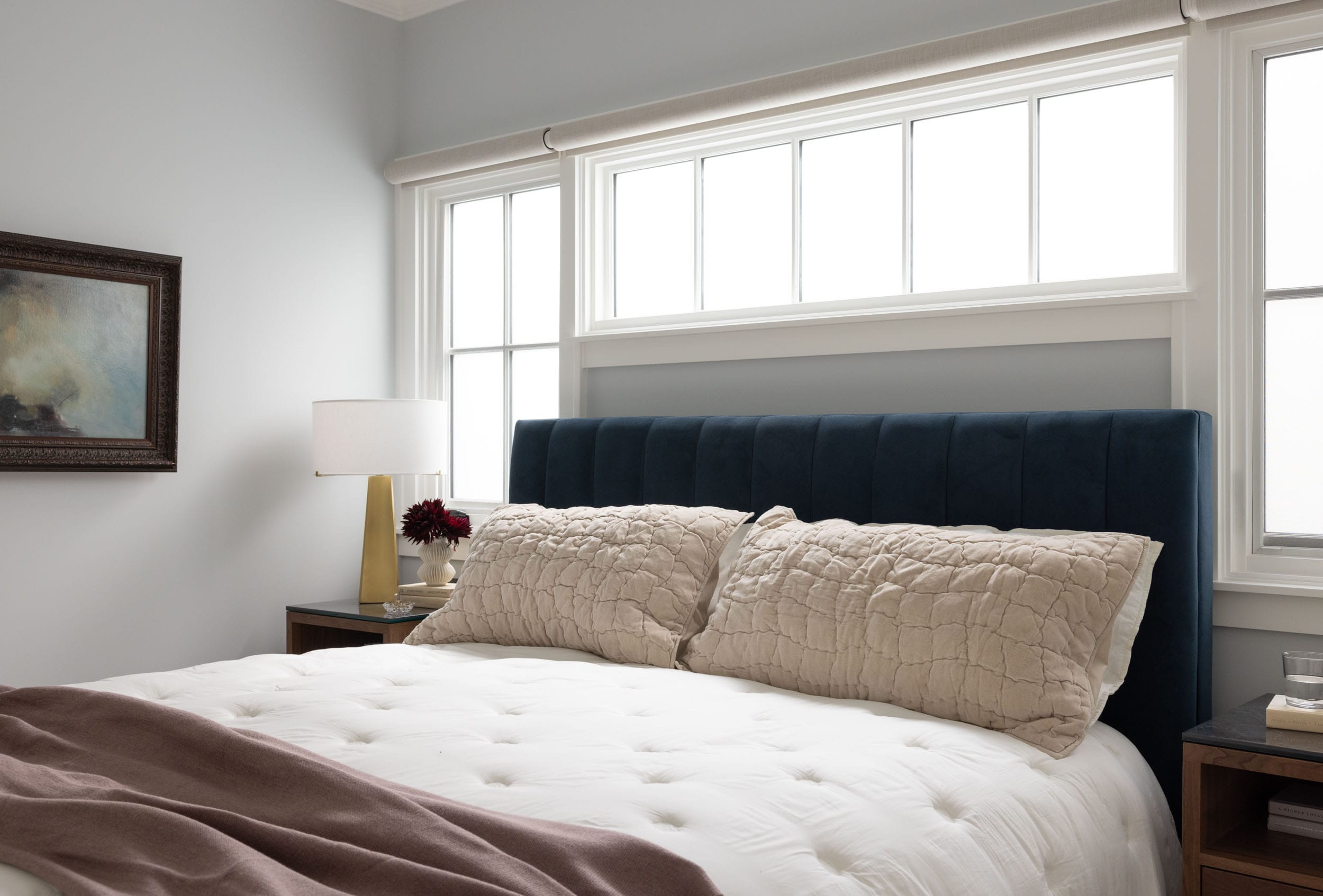 A bedroom with a large bed featuring white bedding and beige pillows, a dark headboard, and side tables with a lamp and flowers. A painting hangs on the light gray wall.