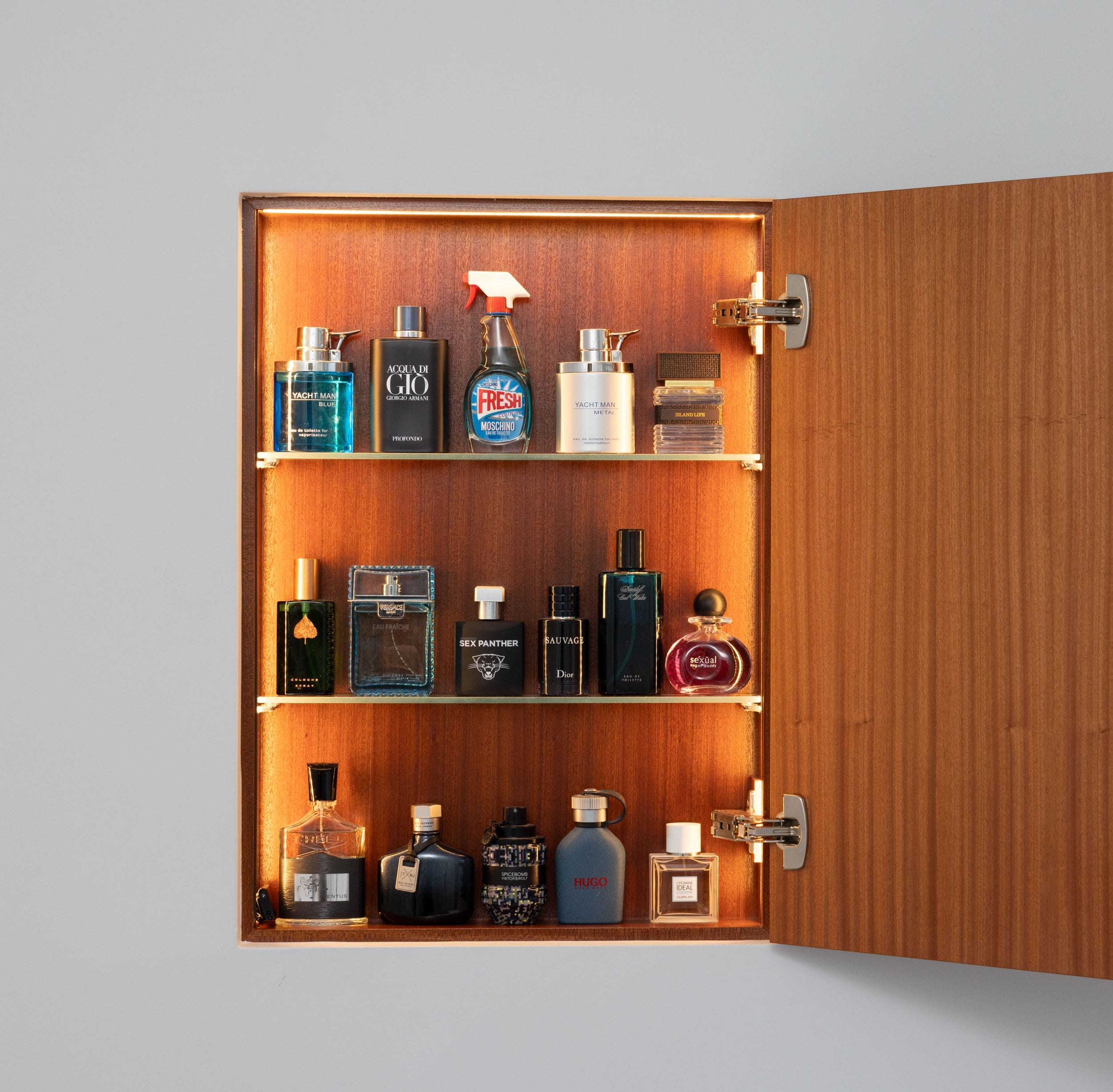 Wooden cabinet open, displaying various bottles of cologne and grooming products arranged on three shelves.