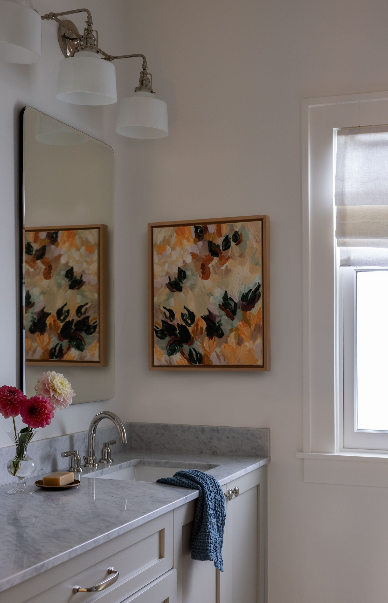 A bathroom with a marble countertop, a sink, and a mirror. A colorful abstract painting hangs on the wall. A towel and a vase with flowers are on the counter. Natural light filters through a window.