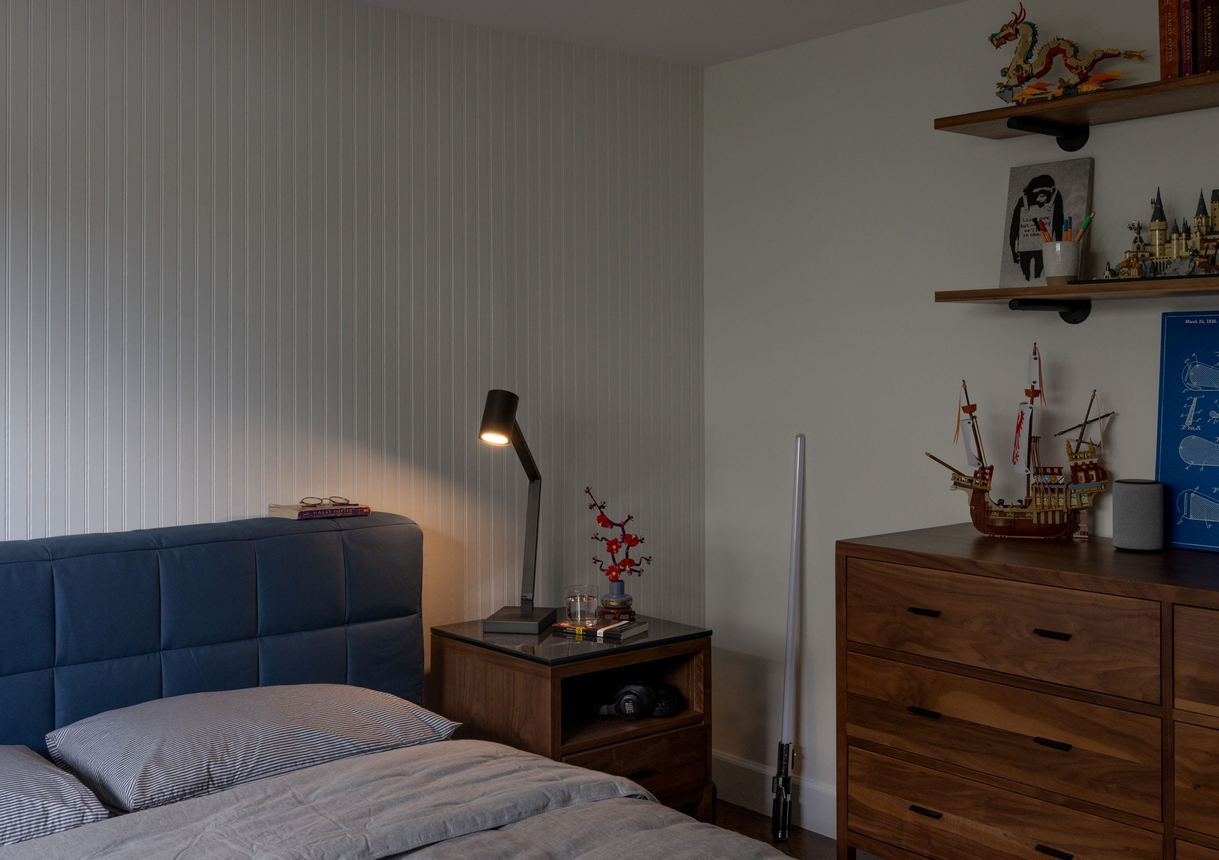 A neatly arranged bedroom with a blue upholstered bed, wooden nightstand, and dresser. Shelves hold various figurines and models. A reading lamp on the nightstand illuminates a molecular structure.