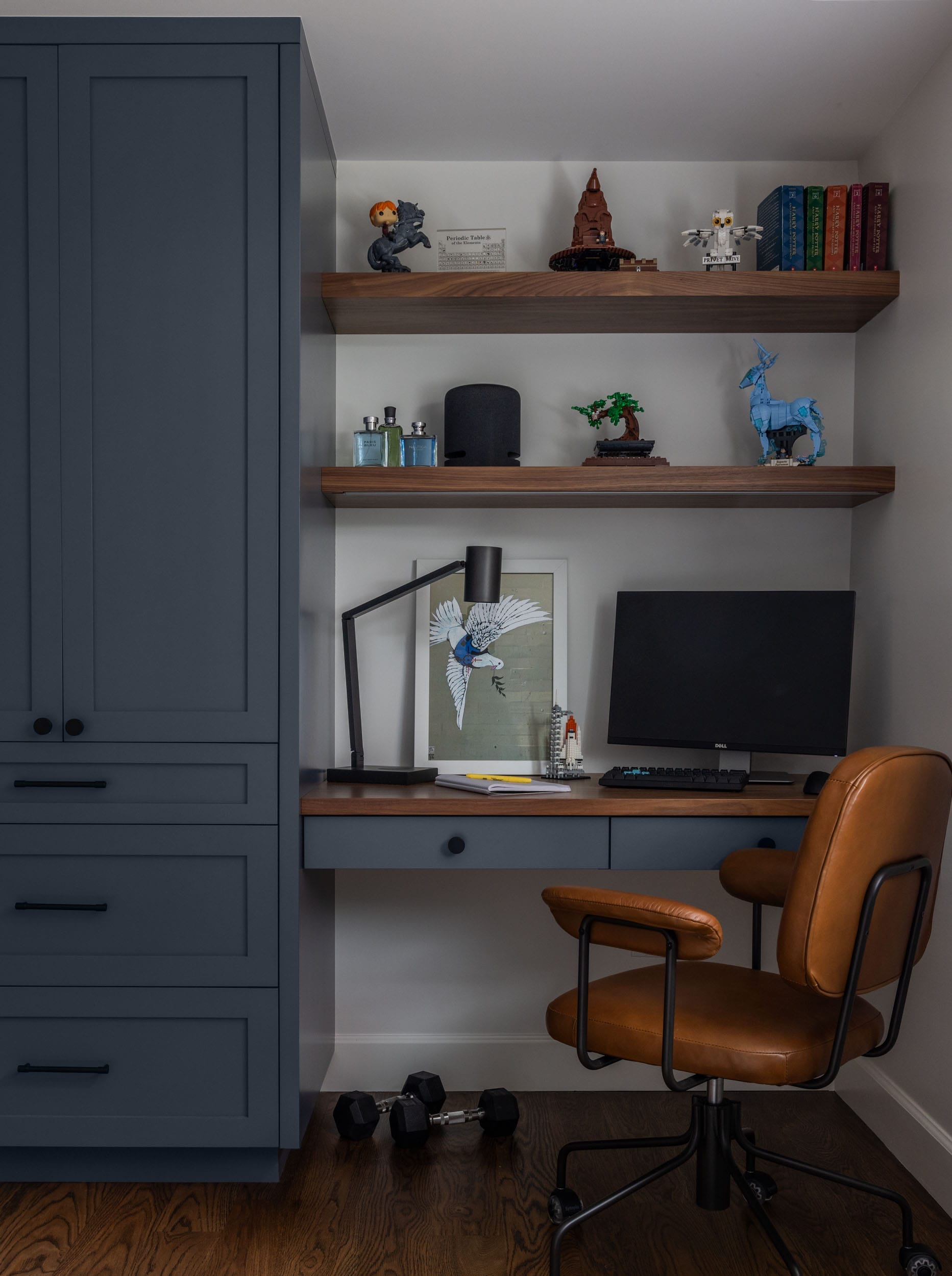 Home office with a wooden desk, brown leather chair, and dark blue cabinet. Shelves hold books, a small tree, and figurines. A computer monitor and a desk lamp are on the desk. Dumbbells on the floor.