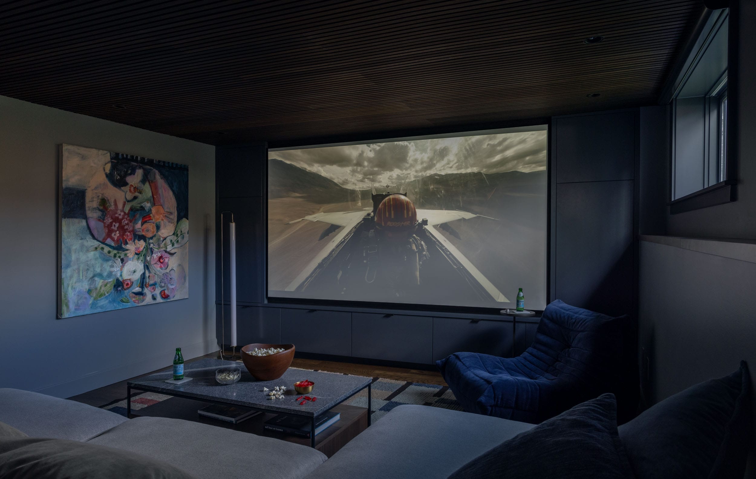 Home theater room with a large screen displaying a jet in flight, a sofa, a blue chair, and a glass table with snacks and drinks. A colorful painting hangs on the left wall.