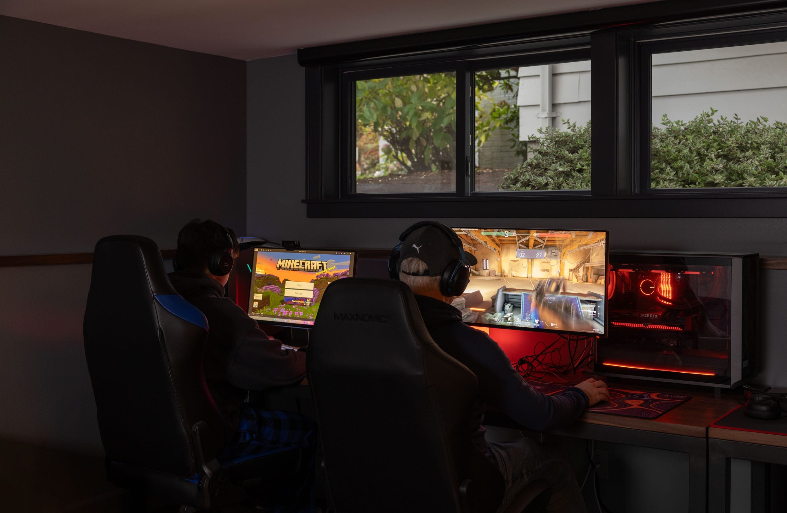 Two people sitting at computer desks, playing video games on separate monitors. One screen shows 