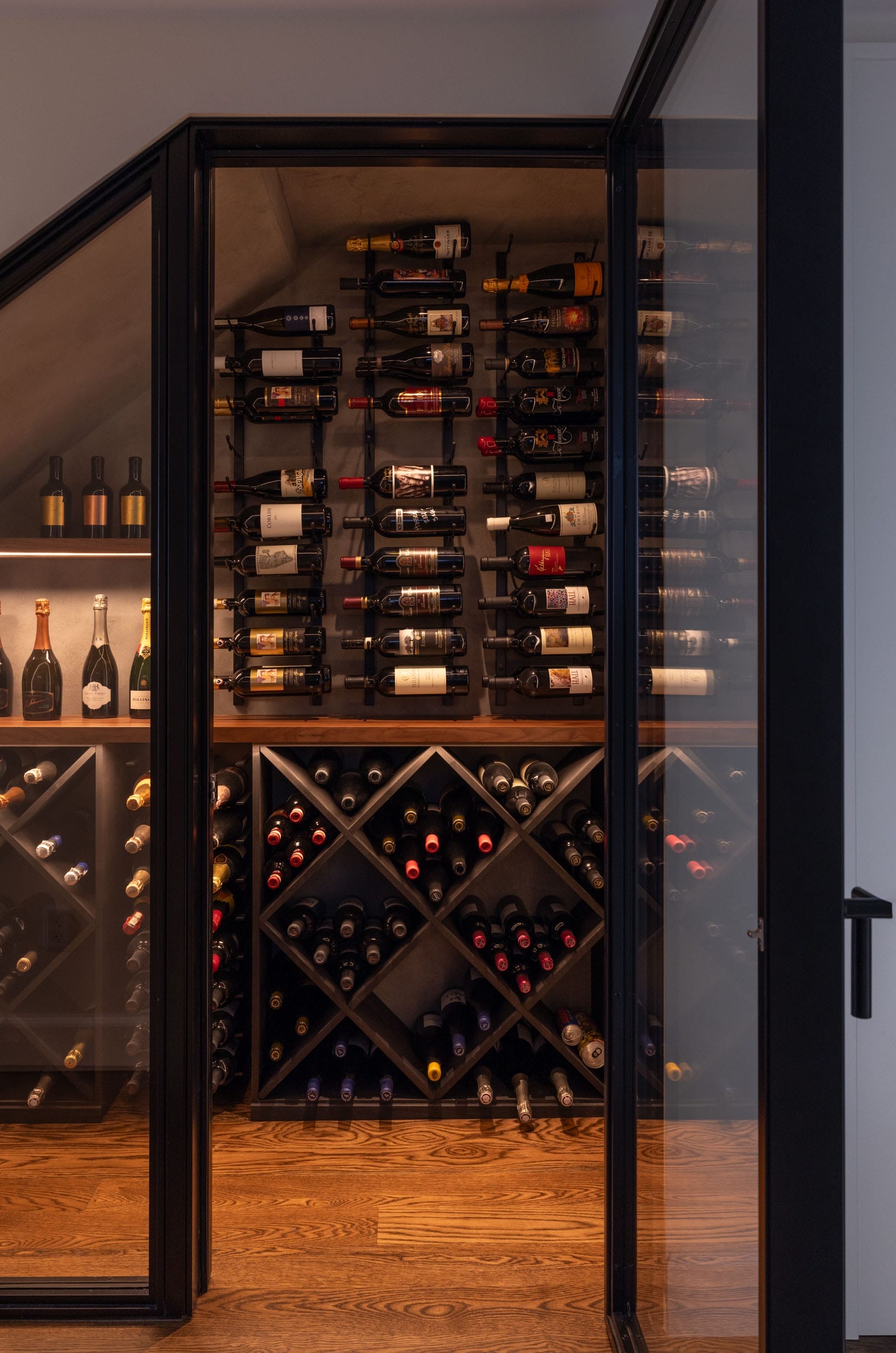 A glass-doored wine cellar with shelves and racks holding various bottles of wine, set against wooden flooring and soft lighting.