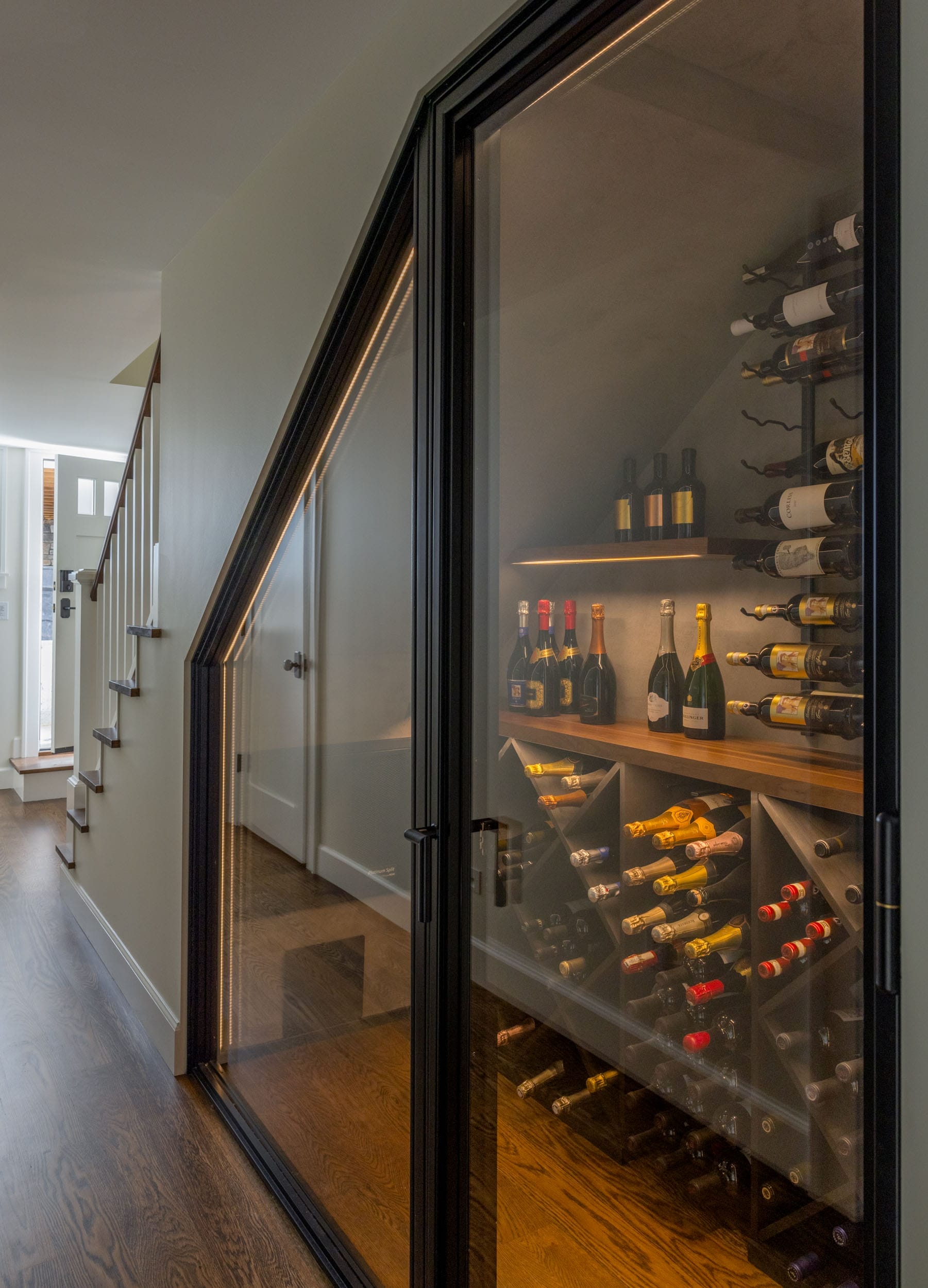 Glass-enclosed wine cellar under a staircase, displaying various wine bottles on wooden shelves and in racks.