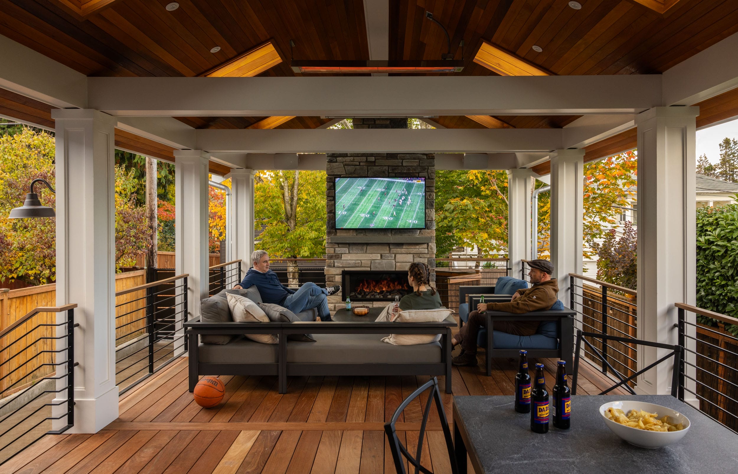 Three people sit on outdoor couches watching a football game on a TV above a fireplace in a covered patio area.