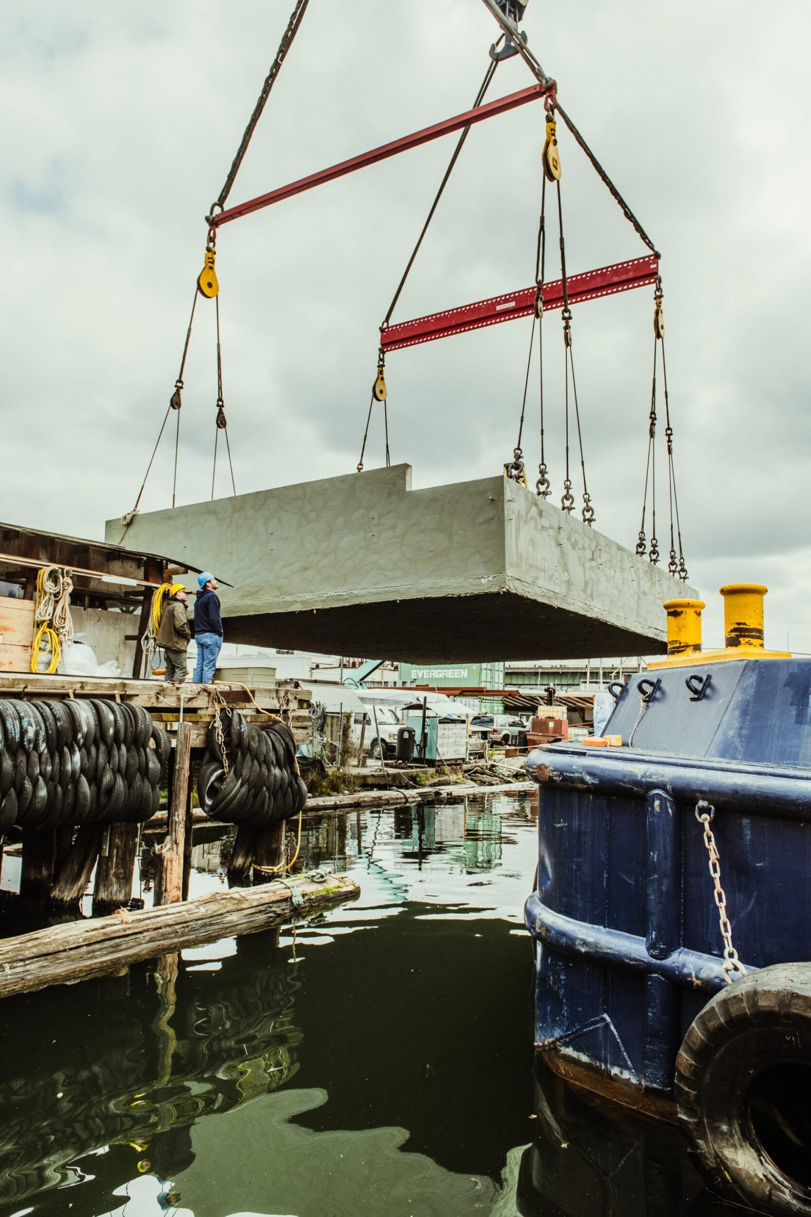 A large concrete block, essential for the ongoing Dyna Contracting project, is being lifted by a crane over water at a dock. Nearby, a worker from Dyna Builders keenly observes the process.