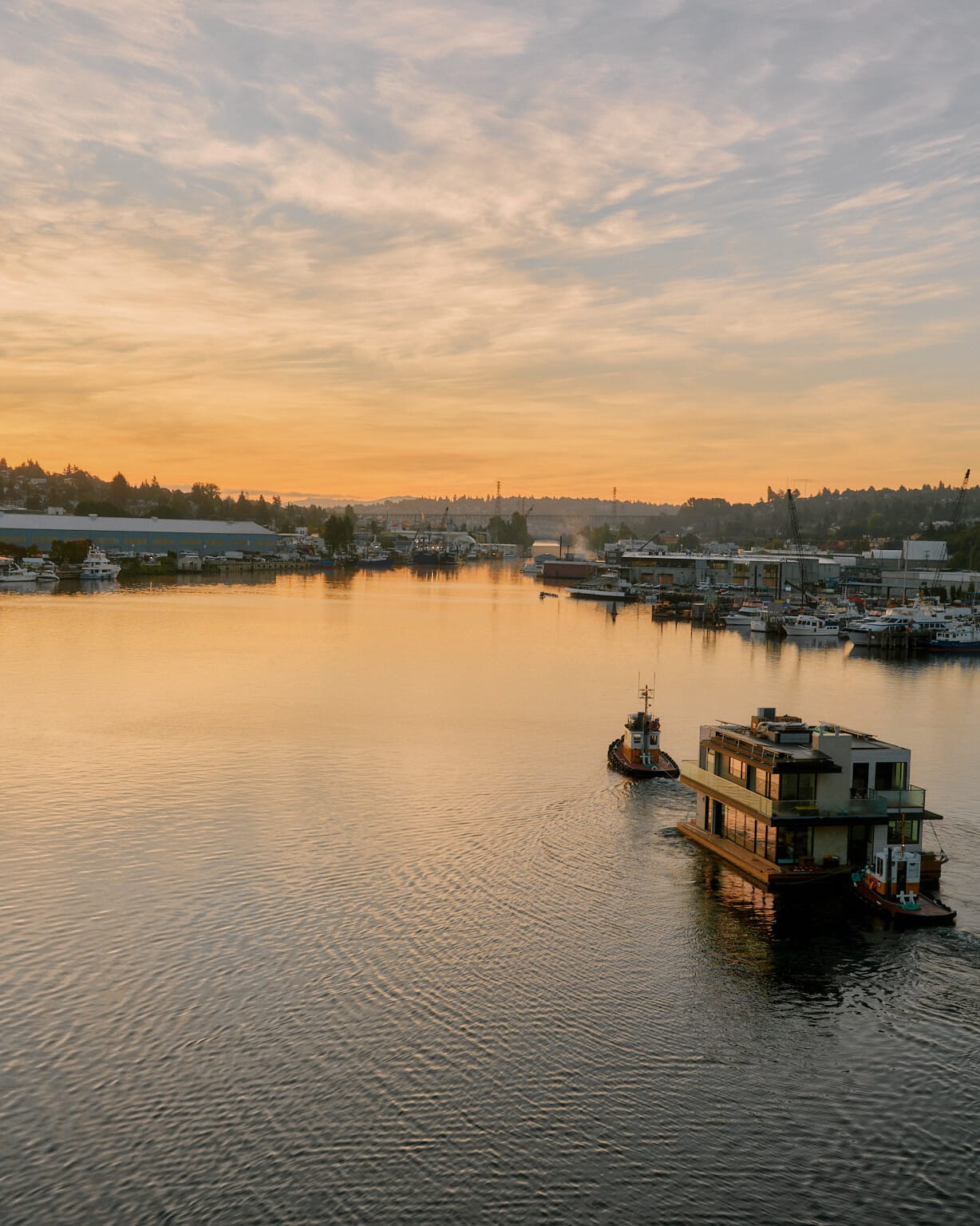 Two boats navigate a calm waterway at sunset, surrounded by docks and distant hills under a partly cloudy sky. Nearby, a Seattle floating homes contractor works on an innovative project, adding to the charm of this serene evening landscape.