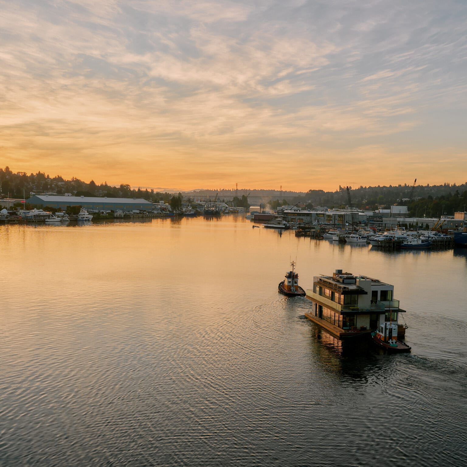 Two boats navigate a calm waterway at sunset, surrounded by docks and distant hills under a partly cloudy sky. Nearby, a Seattle floating homes contractor works on an innovative project, adding to the charm of this serene evening landscape.