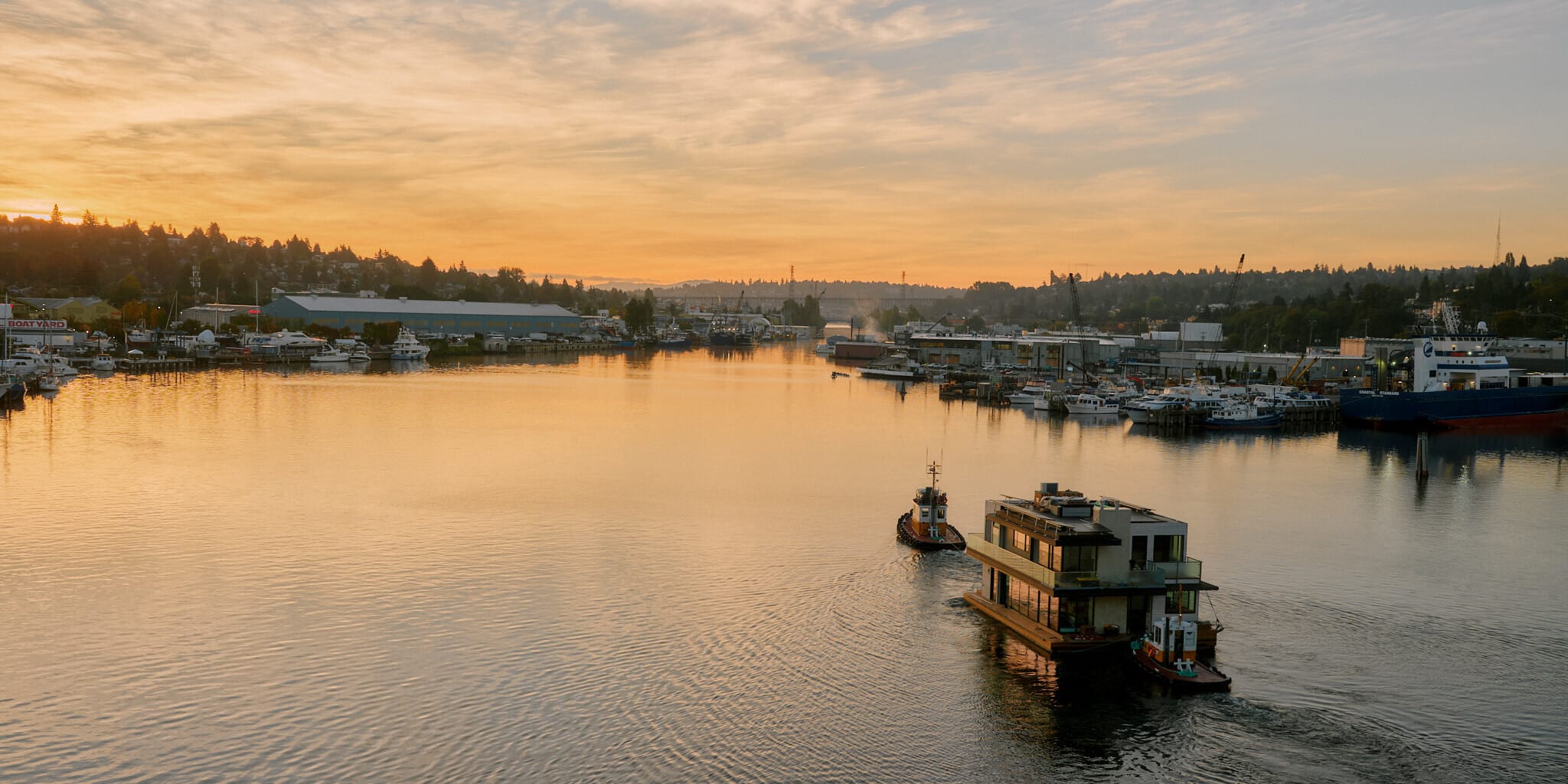 Two boats navigate a calm waterway at sunset, surrounded by docks and distant hills under a partly cloudy sky. Nearby, a Seattle floating homes contractor works on an innovative project, adding to the charm of this serene evening landscape.