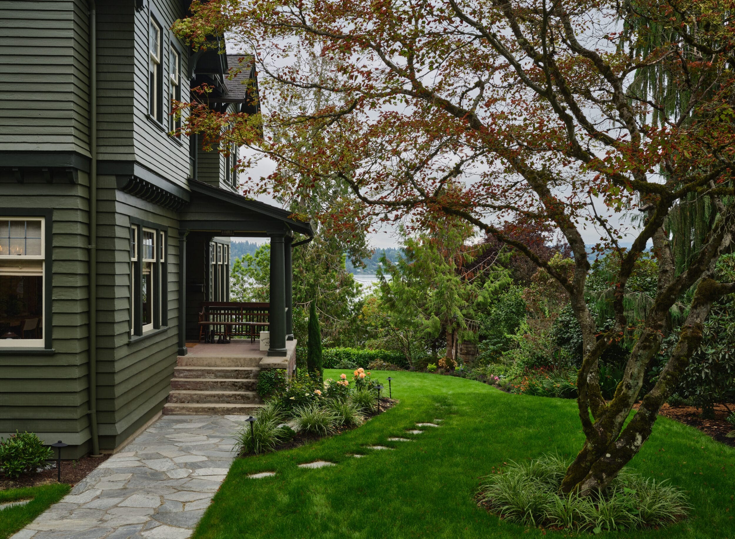 A green house with large windows stands beside a stone path leading through a landscaped garden with a tree and neatly trimmed grass. A distant body of water is visible through the trees.