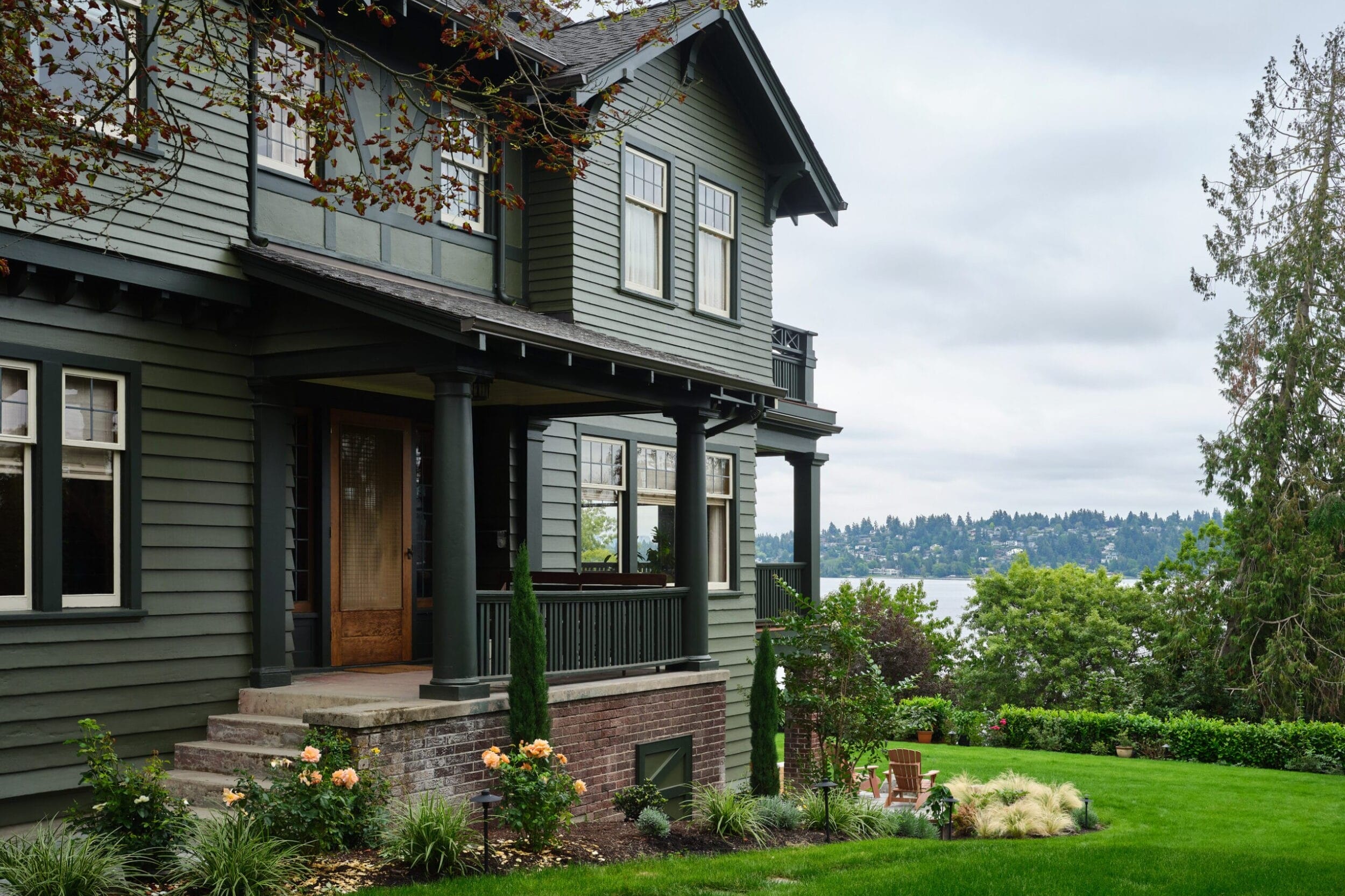 A green two-story house with a porch sits in a landscaped yard overlooking water and distant hills.